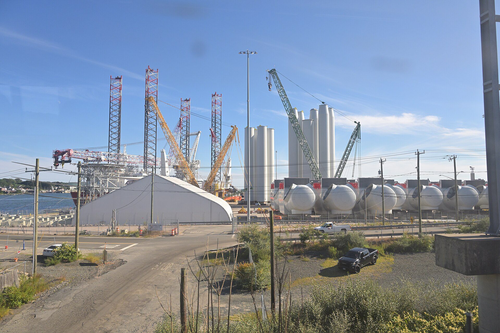 Upright white wind turbine tower sections and a row of nacelles staged at a waterfront port, with the legs of a jack-up vessel and several cranes behind them