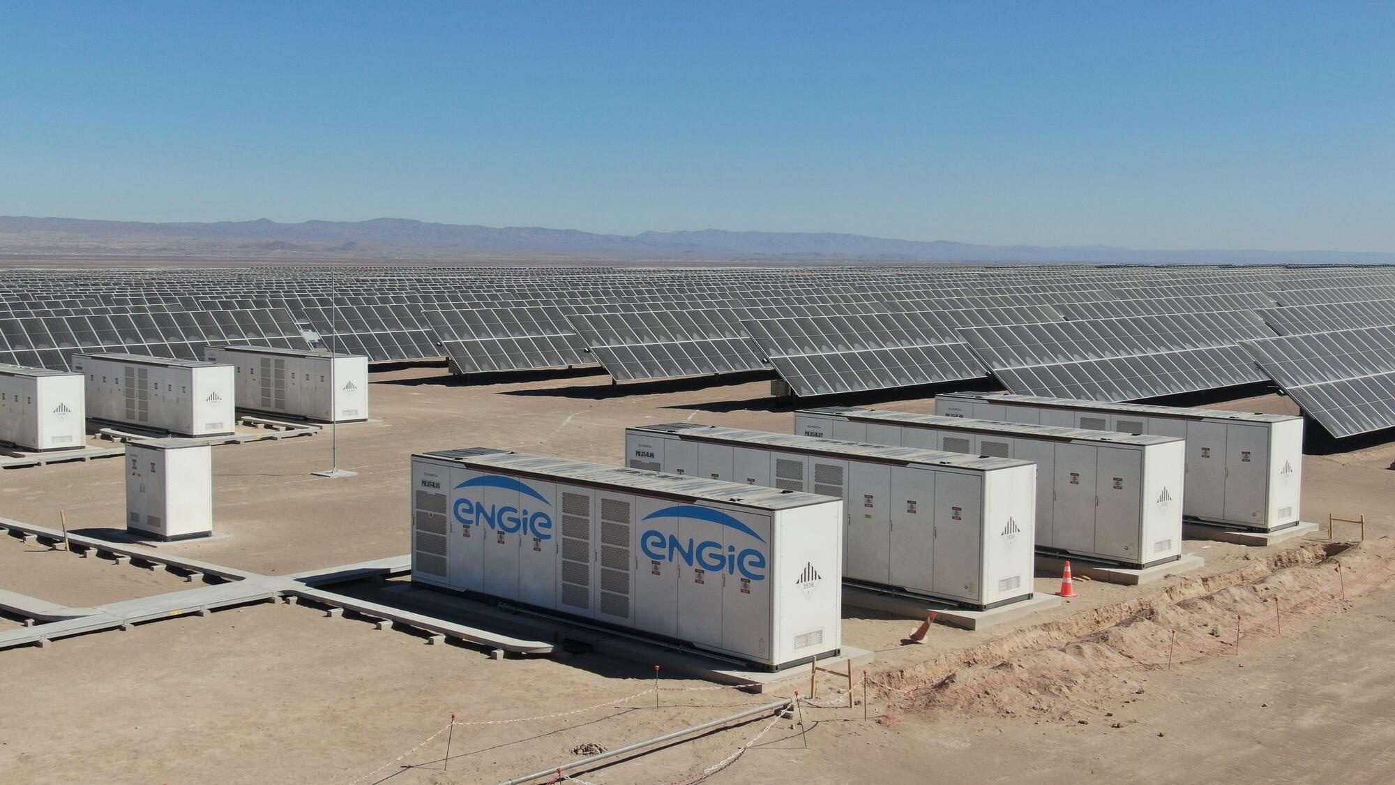 ENGIE-branded battery storage containers on concrete pads in a sandy yard, with long rows of solar panels and distant mountains behind under a clear sky