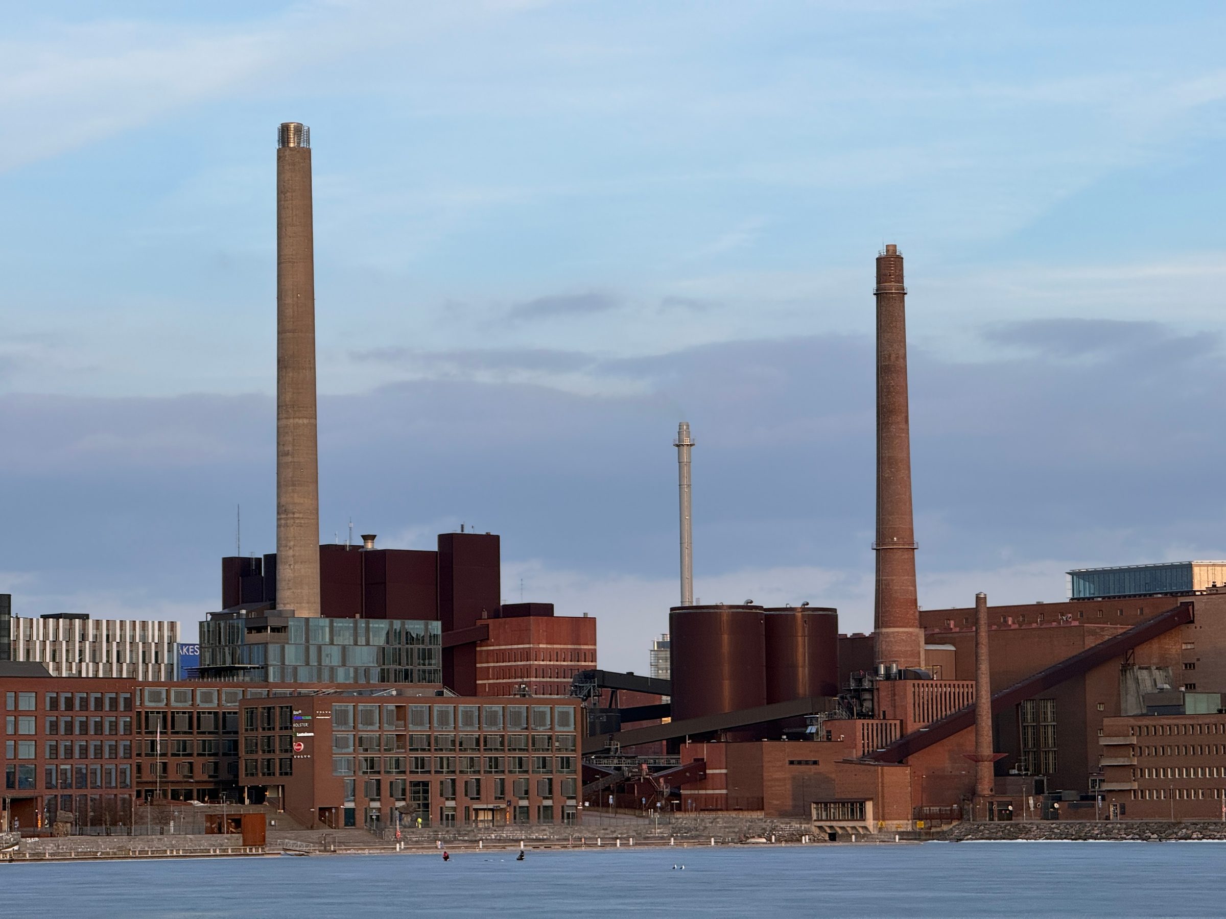 The Salmisaari power plant complex on the Helsinki waterfront in March 2026: a tall concrete stack, a slim metal stack, a tall brick stack and a shorter brick chimney above dark brick plant buildings and fuel silos, seen across open water.