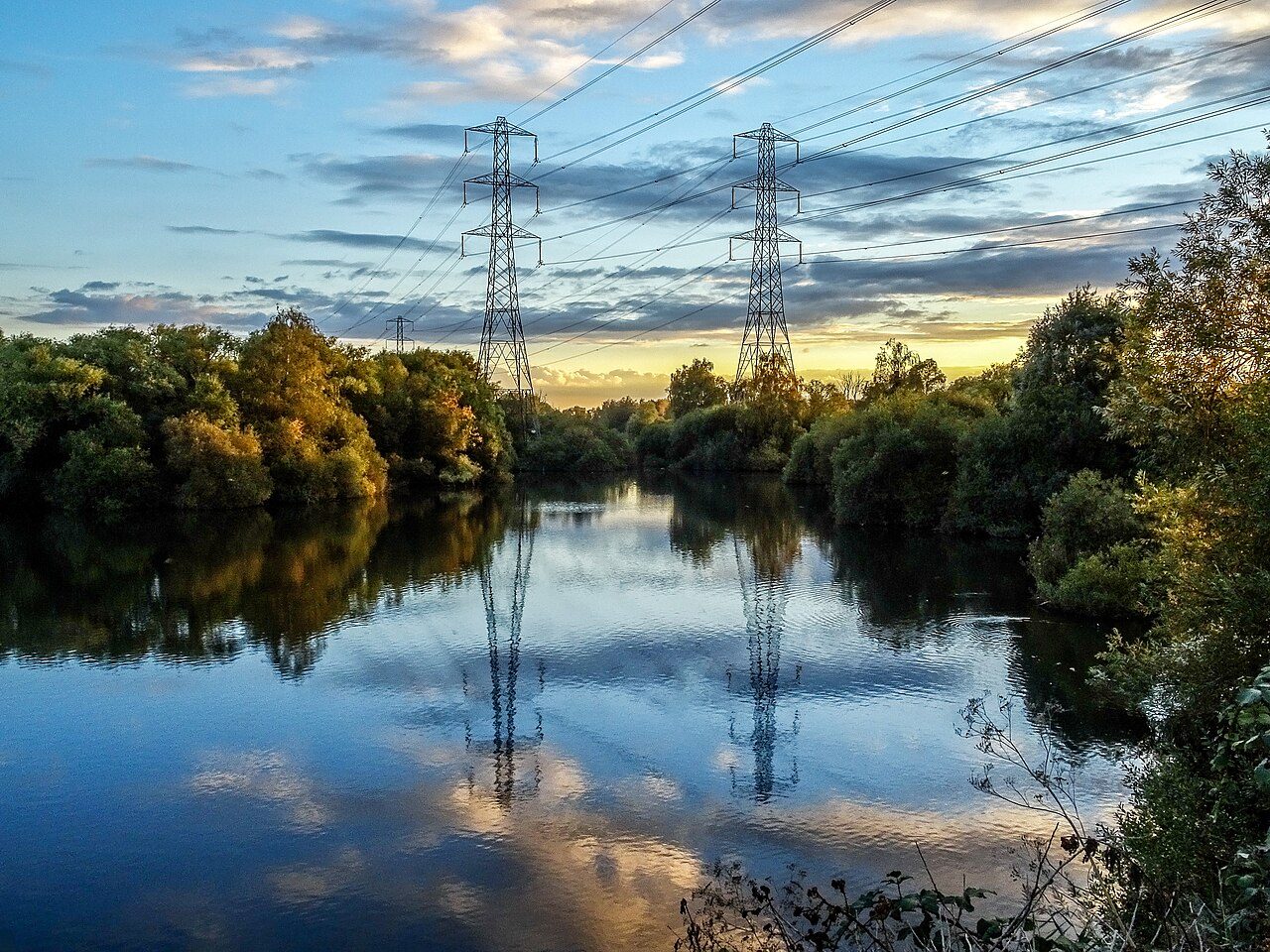 Two steel lattice pylons carrying high-voltage lines over a still wetland lake in the Lea Valley, England, with trees on both banks, a low sun lighting the clouds and both pylons reflected in the water.
