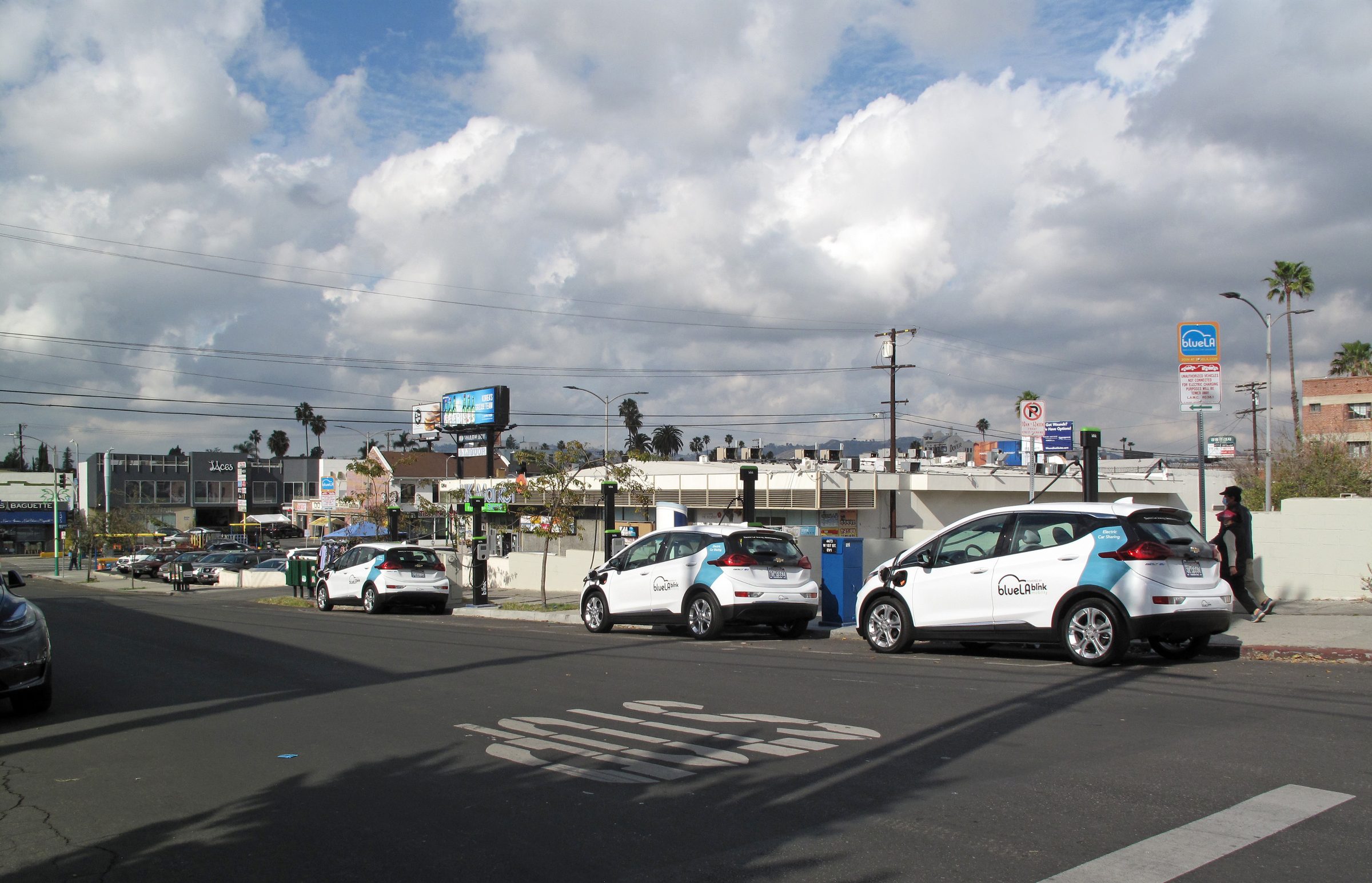Three white BlueLA car-share Chevrolet Bolts at blink curbside charging posts on a Koreatown street in Los Angeles, the two nearest with charging cables plugged into their front fenders, under a partly cloudy sky