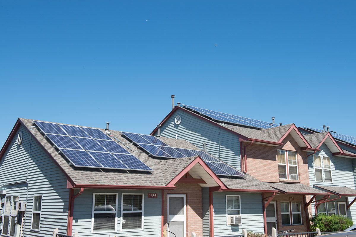 Solar panel arrays on the gray shingle roofs of a row of two-story townhomes with blue-gray siding and brick fronts under a clear blue sky in Denver, Colorado