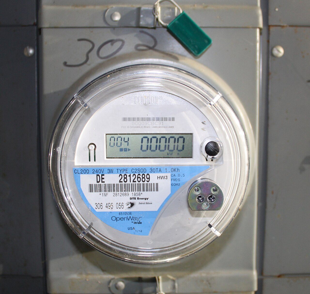 A round residential electricity meter with a DTE Energy label and a digital kWh display, mounted on a gray metal box with a green tag on its latch