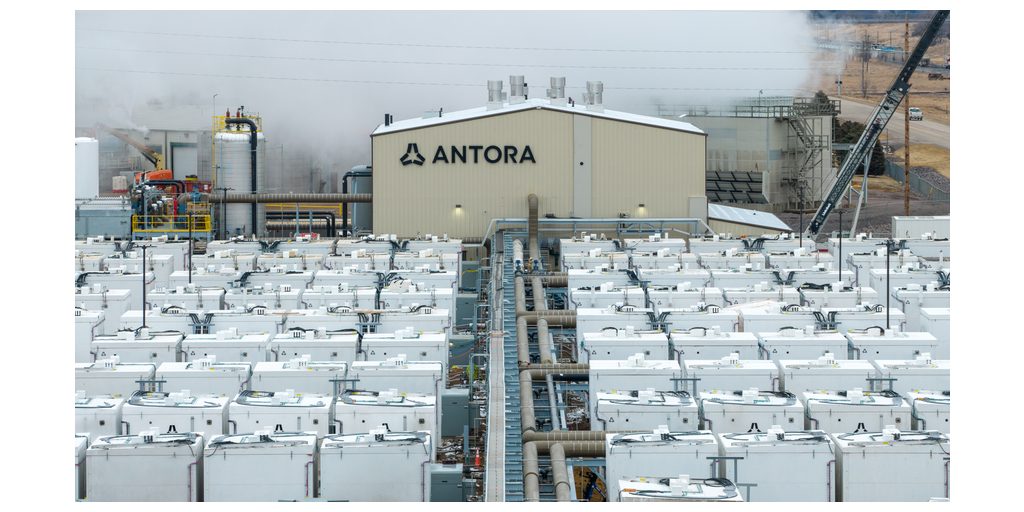 Elevated view of rows of white Antora thermal battery enclosures with a pipe rack running between them toward a beige building marked ANTORA, white tanks at left, a crane at upper right and white plumes rising behind the building at Antora's Big Stone City, South Dakota project