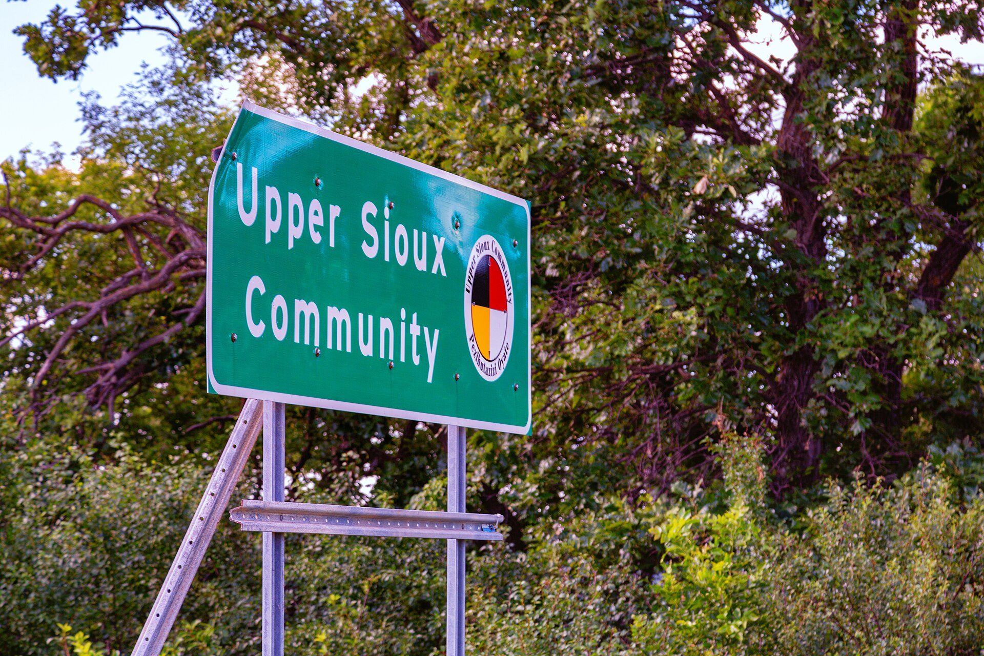 Green highway sign reading Upper Sioux Community with the community's round seal, mounted on two metal posts in front of leafy trees near Granite Falls, Minnesota