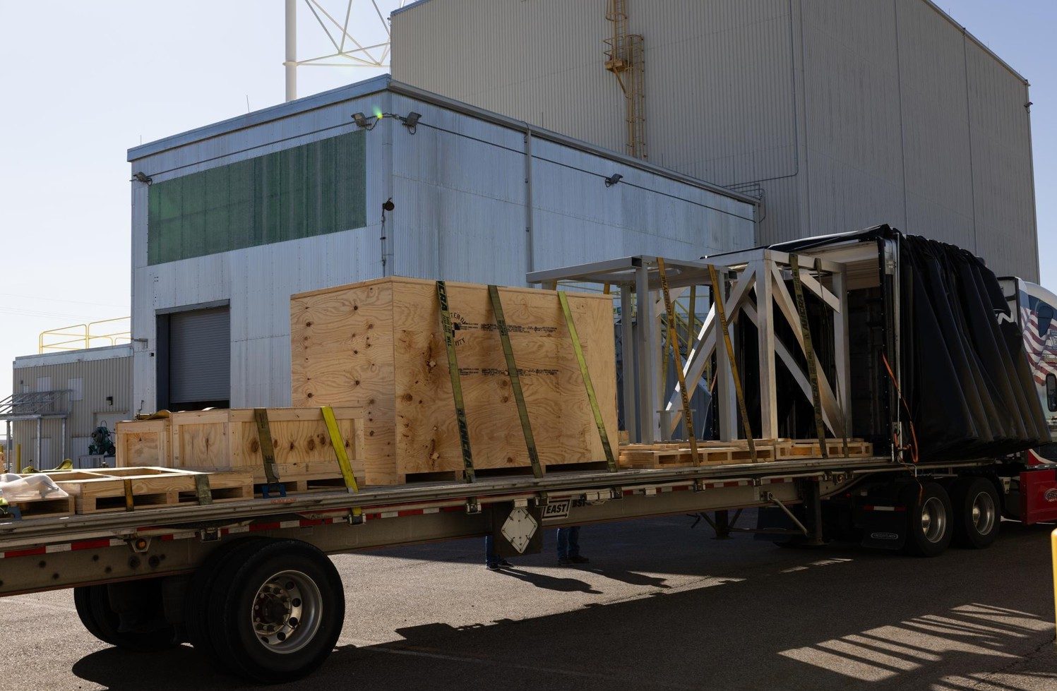 A flatbed trailer loaded with strapped wooden shipping crates and white metal frames, parked beside tall corrugated metal buildings under a clear sky