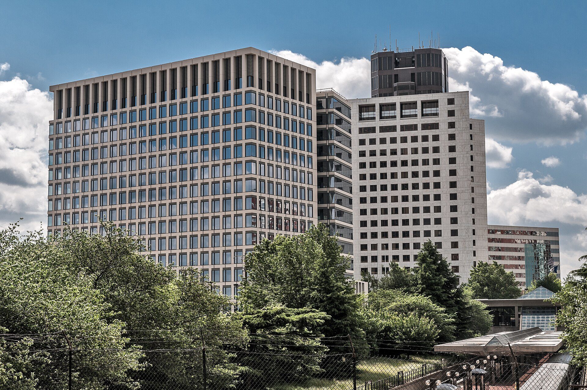 Office towers of the NRC headquarters campus in Rockville, Maryland, rising above trees, with a Metro station canopy and a train at lower right.