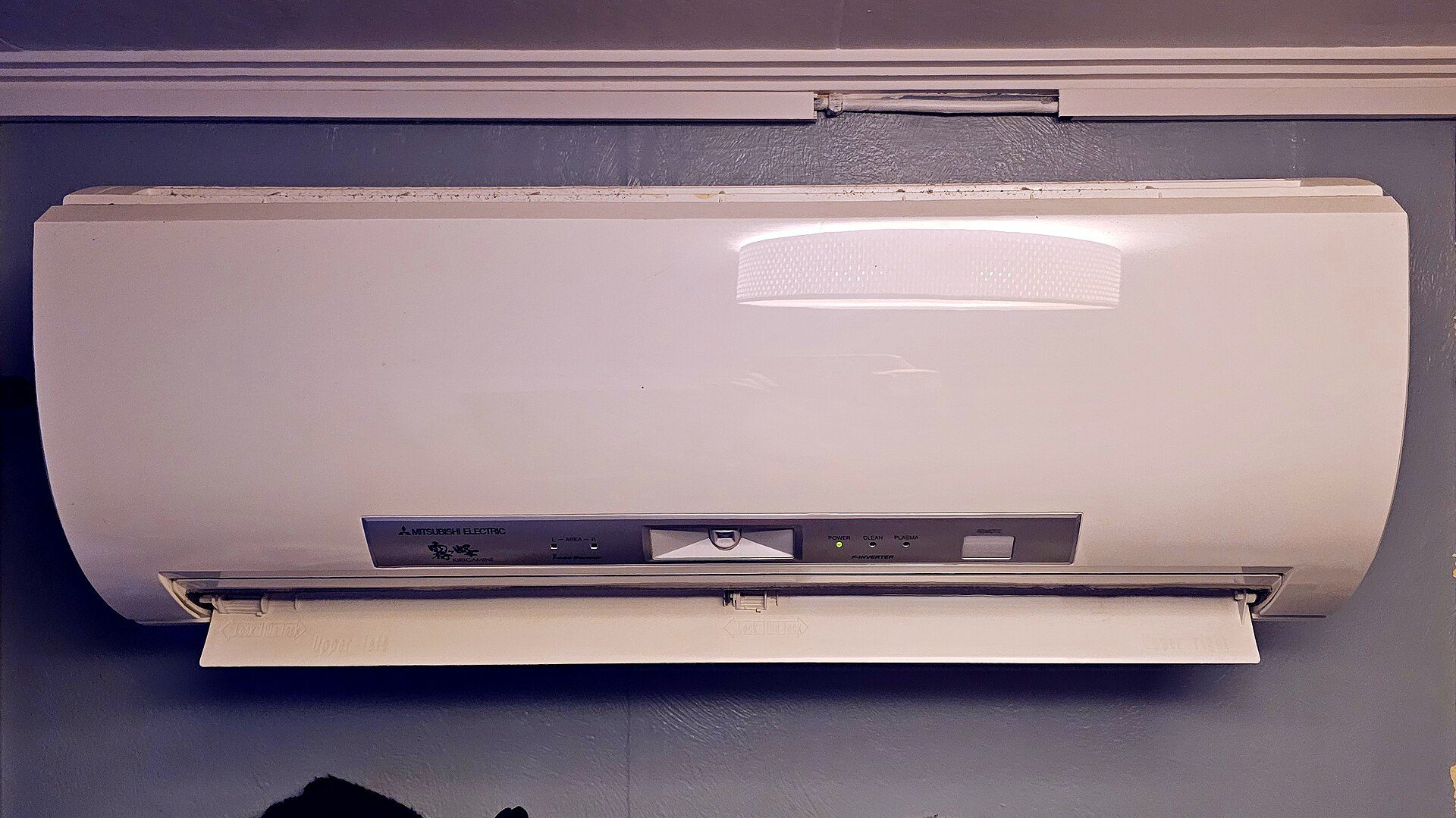 White wall-mounted indoor unit of a ductless heat pump below a ceiling, lit from above, with its louver open and its control panel visible.