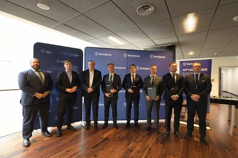 Eight men in dark suits stand in a row in front of Polskie Elektrownie Jądrowe and Westinghouse backdrops, four of them holding dark folders, beside an AP1000 banner.