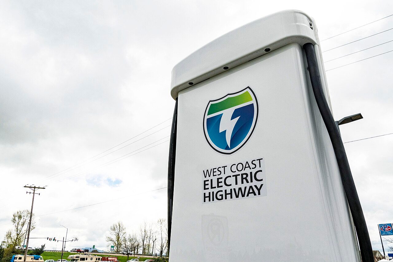 A white charger cabinet bearing a West Coast Electric Highway shield logo, with black cables down its side, photographed from below against an overcast sky with power lines and a road behind it.