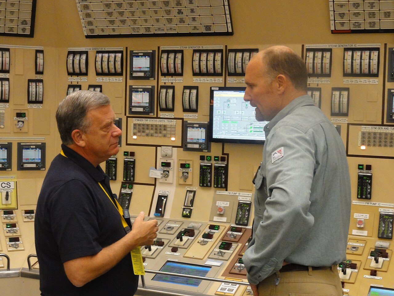 Two men talk in a nuclear plant control room in front of panels of gauges and switches labeled for steam generators and feedwater pumps