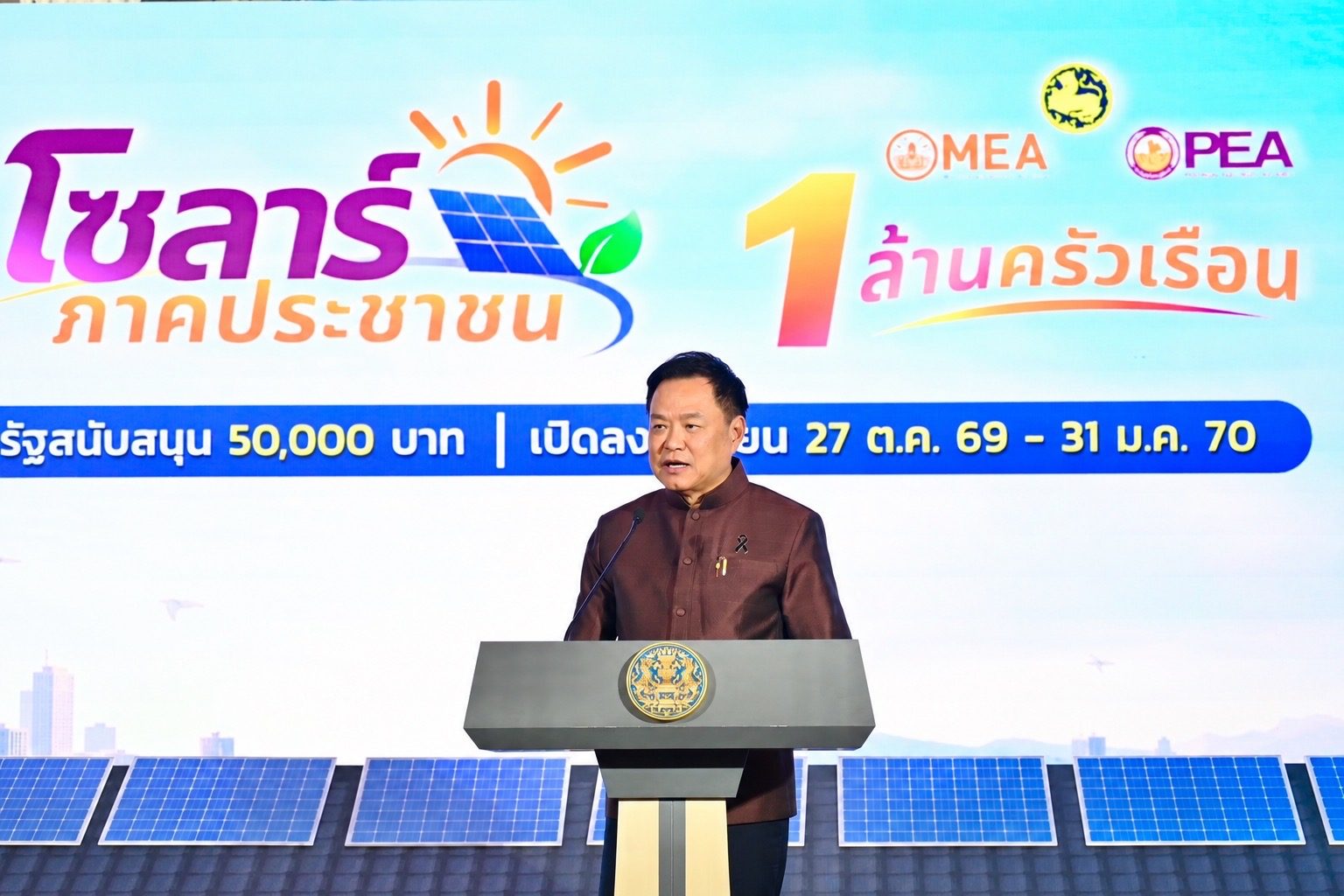 A speaker at a lectern bearing the Thai government emblem, in front of a backdrop advertising People's Solar for 1 million households, 50,000 baht of state support and registration from October 27, 2026 to January 31, 2027