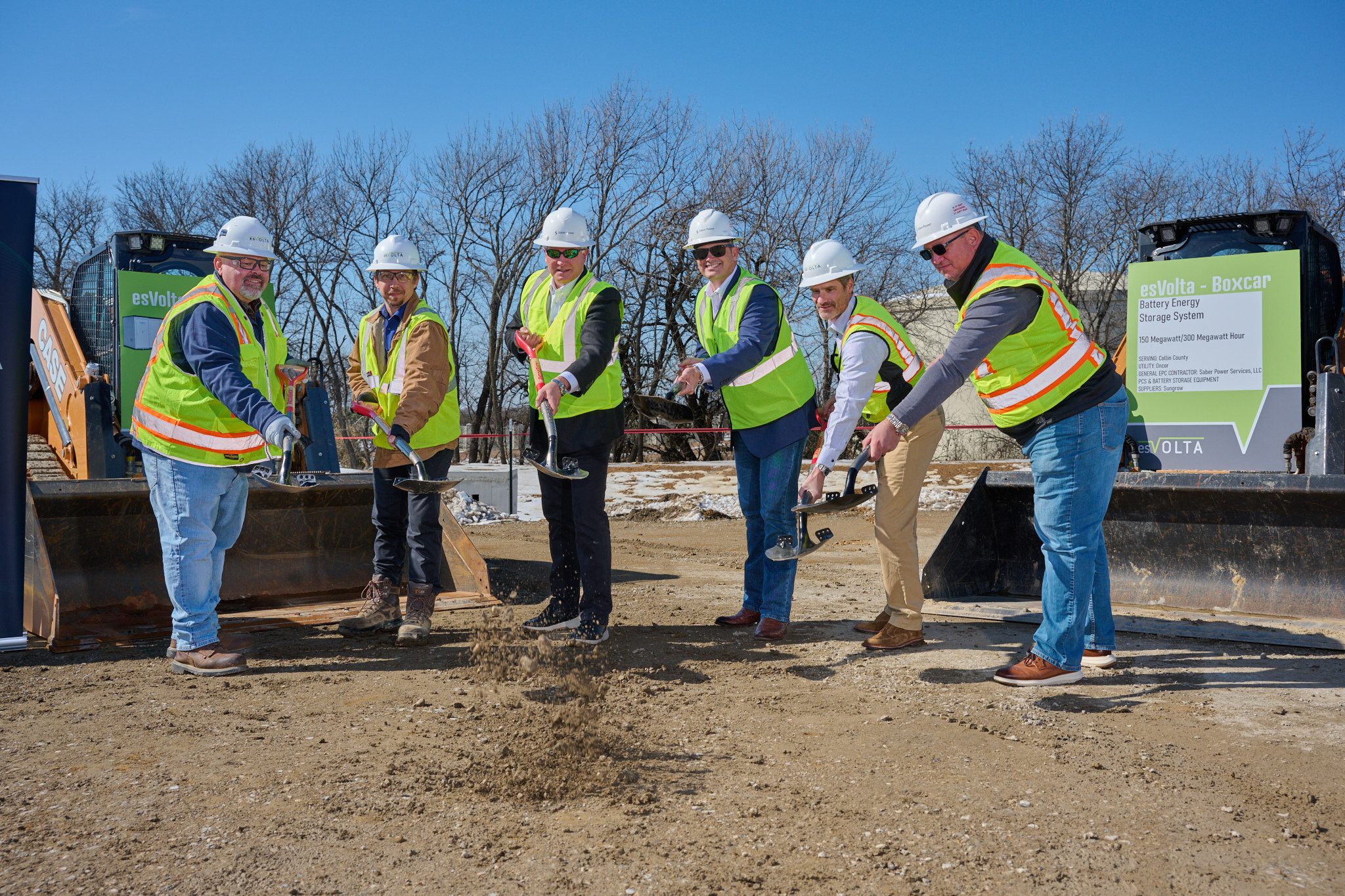 Six men in white hard hats and high-visibility vests toss shovelfuls of dirt between two loader buckets at esVolta's Boxcar groundbreaking, with a light-green sign at right headed esVolta - Boxcar in white lettering above a white inset panel, and bare trees behind