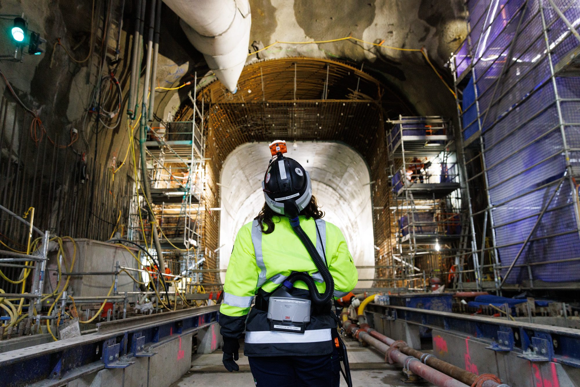A worker in a high-visibility jacket, helmet and powered respirator walks along rails toward a concrete tunnel opening framed by steel reinforcing mesh and scaffolding.