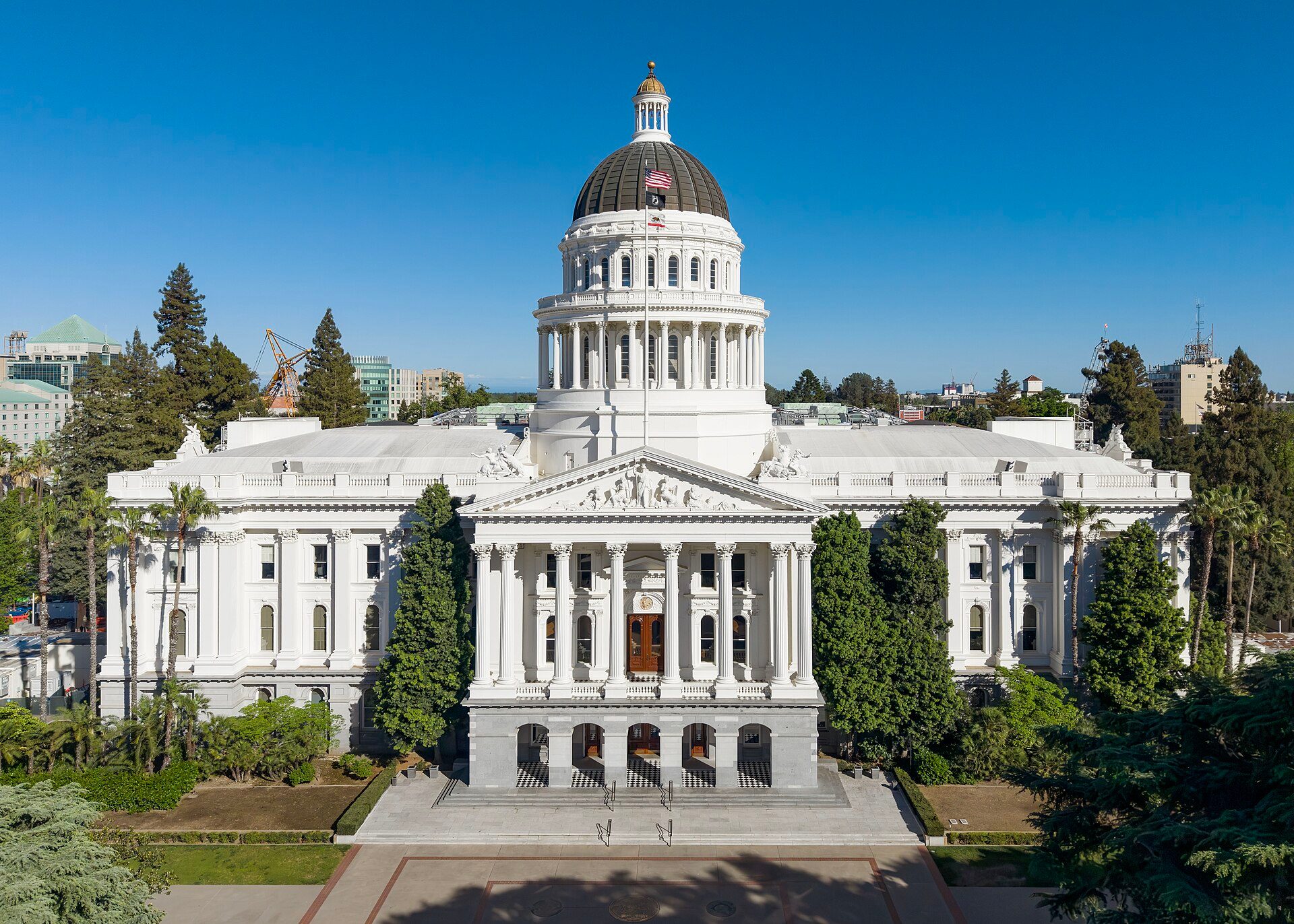 The California State Capitol in Sacramento: white columned facade and drum below a dark dome with a gold cupola, flags on the mast in front, trees on both sides under a clear blue sky