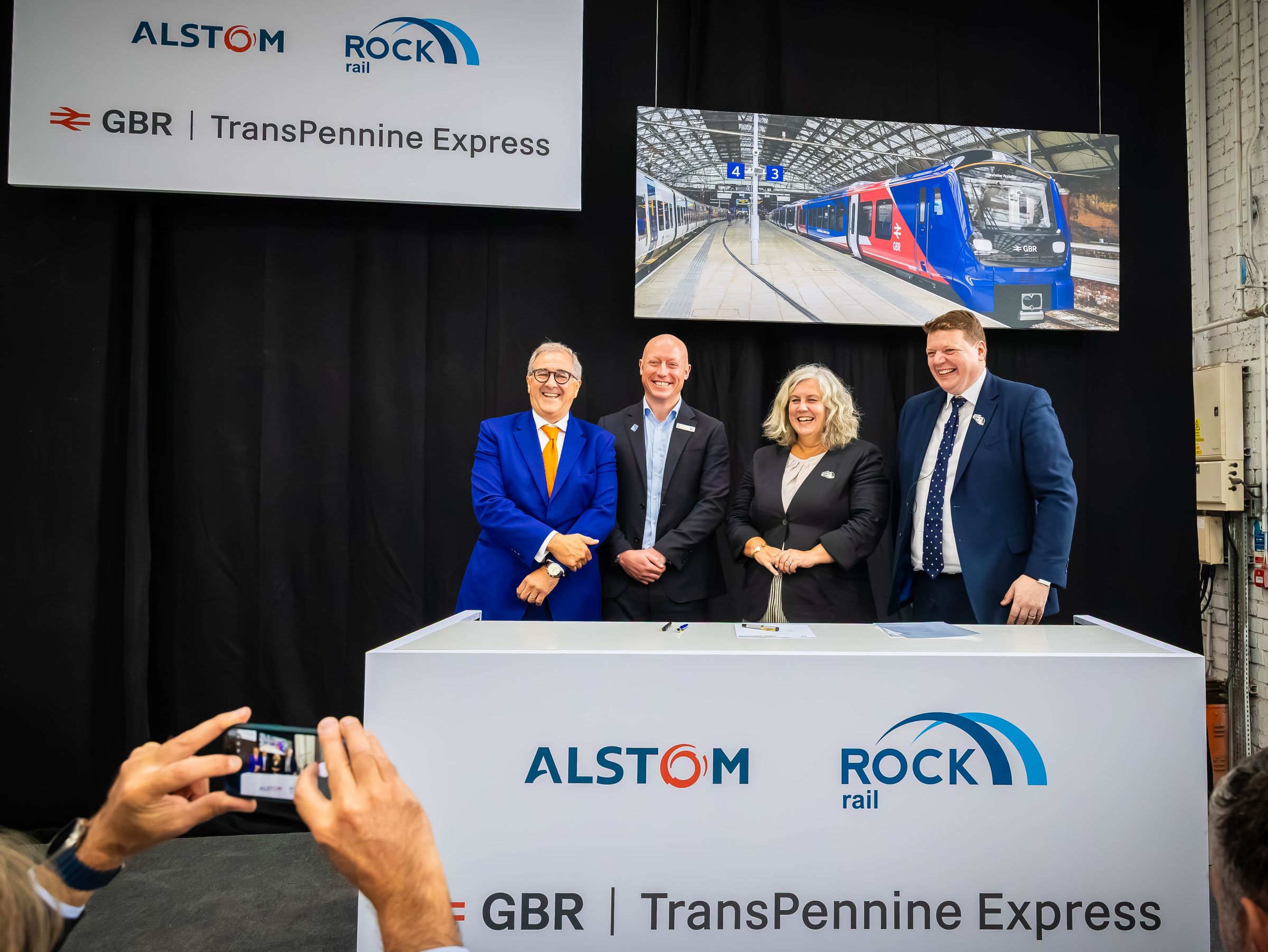 Four people stand behind a white signing desk carrying Alstom and Rock Rail logos, in front of a black curtain with a rendering of a blue and red train on a panel above them