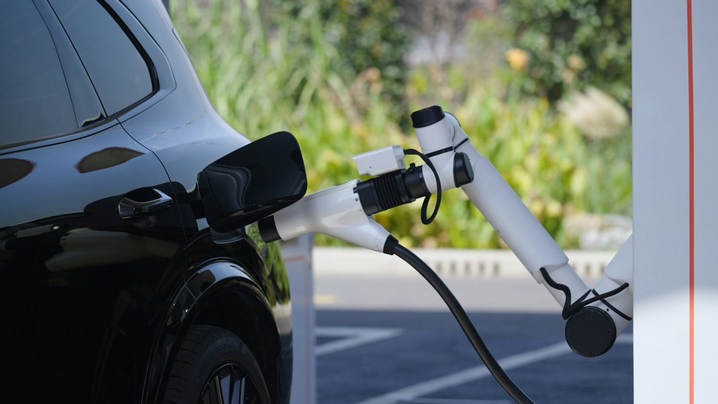 A white robotic charging arm from a Geely charging station plugged into the open charge port of a black car, with green shrubs and a paved lot behind it