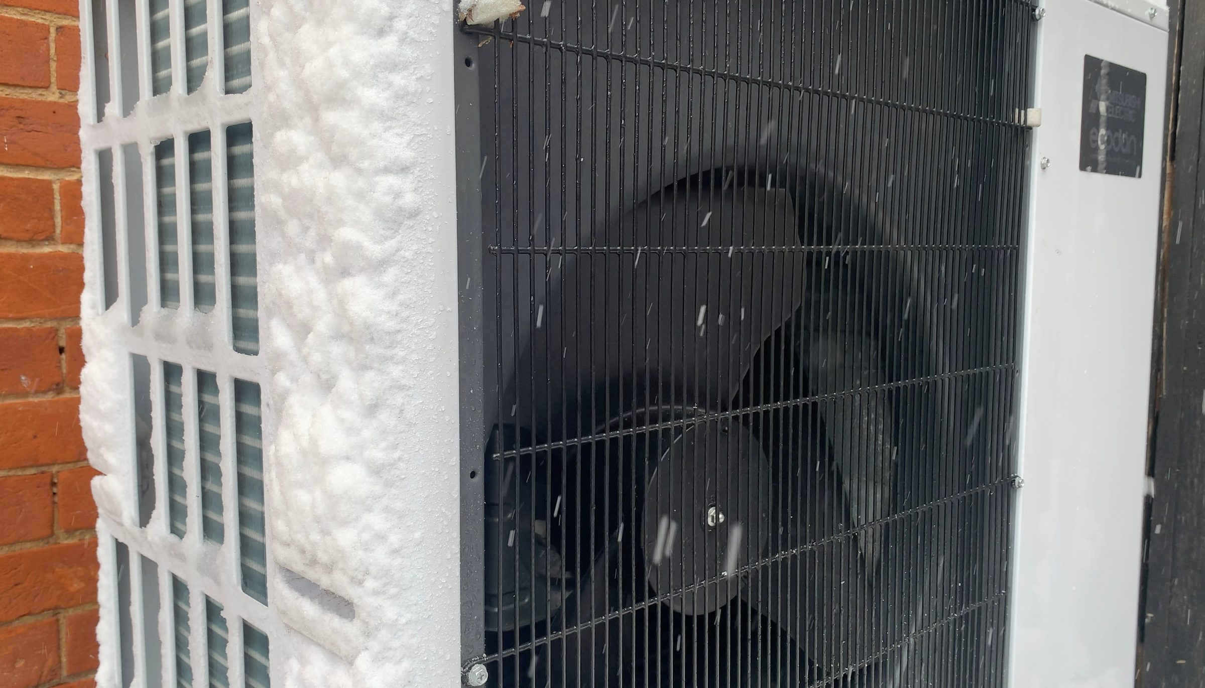 Close-up of a Mitsubishi Electric Ecodan air-source heat pump outdoor unit beside a brick wall, its side grille and coil fins crusted with snow while the front fan grille stays clear and snow falls