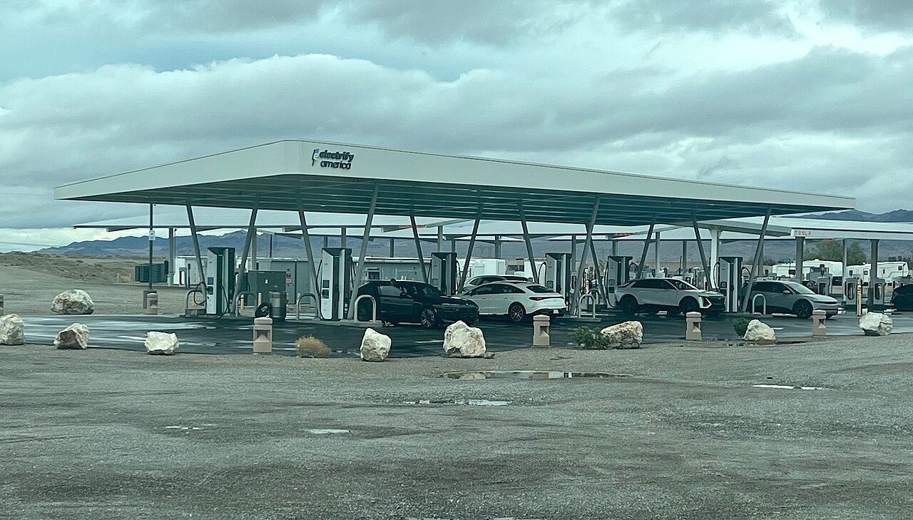 Cars parked at fast chargers under a white Electrify America canopy on a gravel lot, with mountains and a second canopy behind
