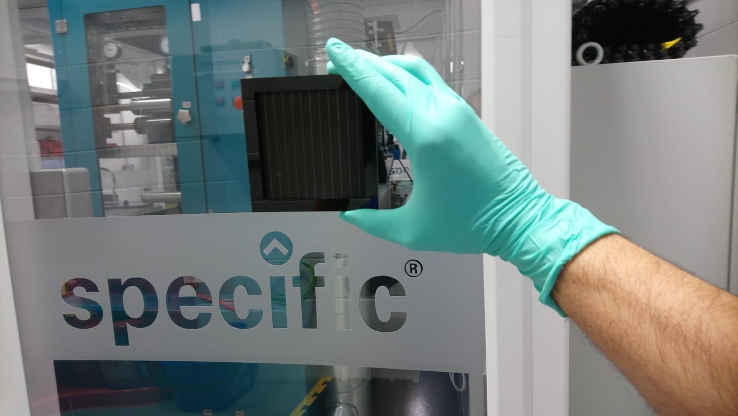 Gloved hand holding a small dark square perovskite solar module in front of laboratory equipment behind glass lettered specific