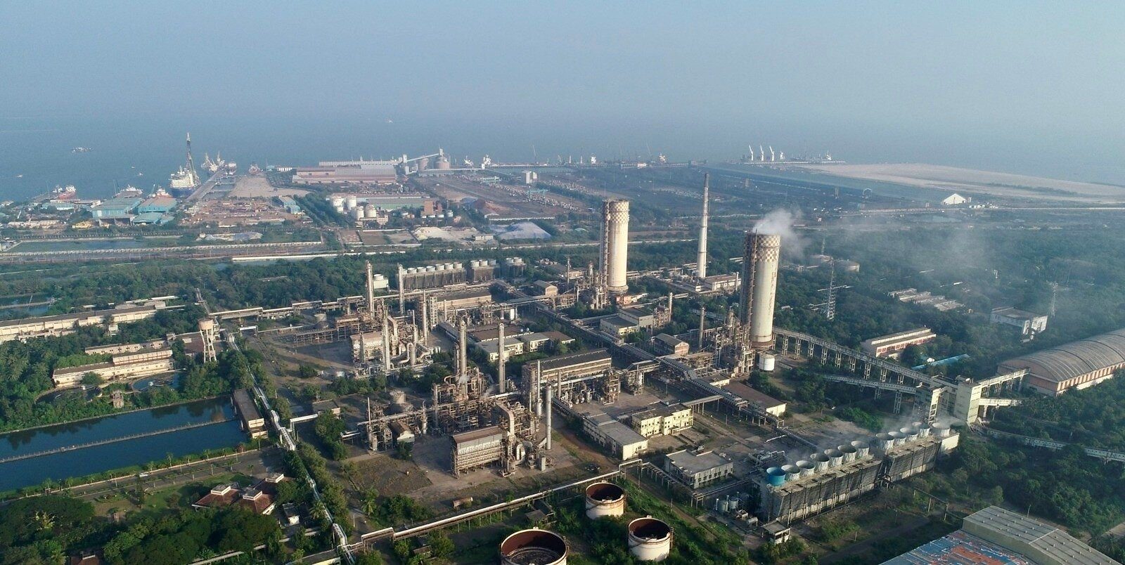 Aerial view of a fertilizer complex with two tall towers, one trailing a white plume, beside process units, with a seaport, docked ships and cranes beyond it under haze