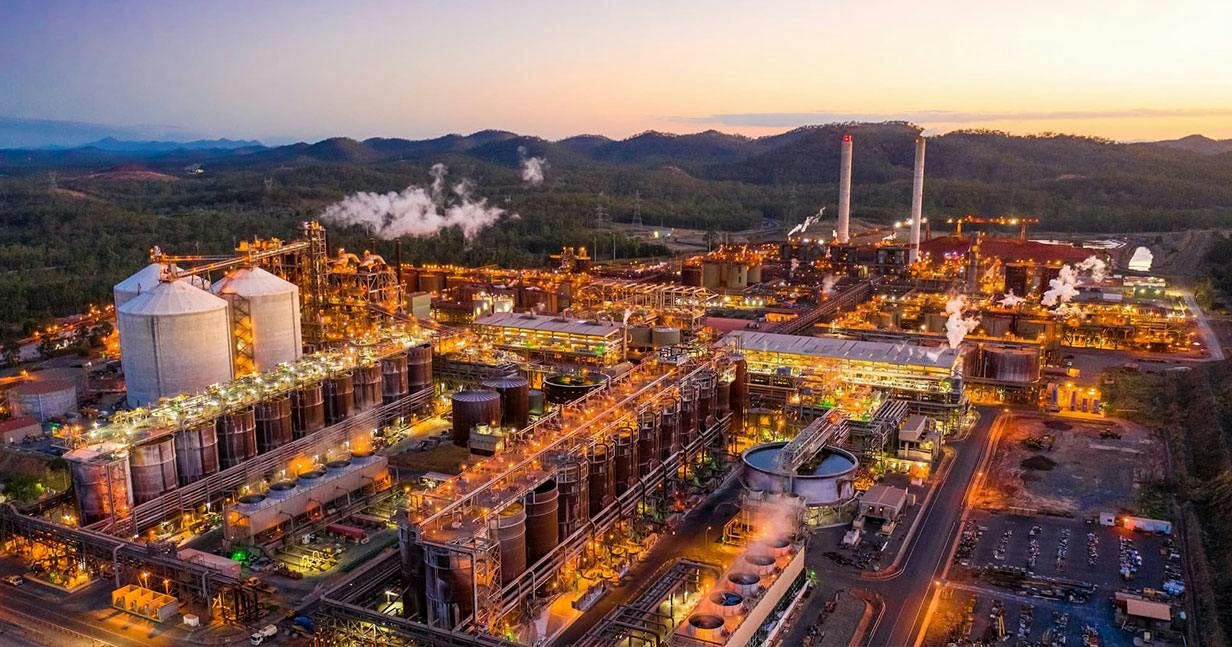 Aerial view at dusk of a brightly lit alumina refinery with white silos, rows of tanks, two tall stacks and steam plumes, with wooded hills behind.