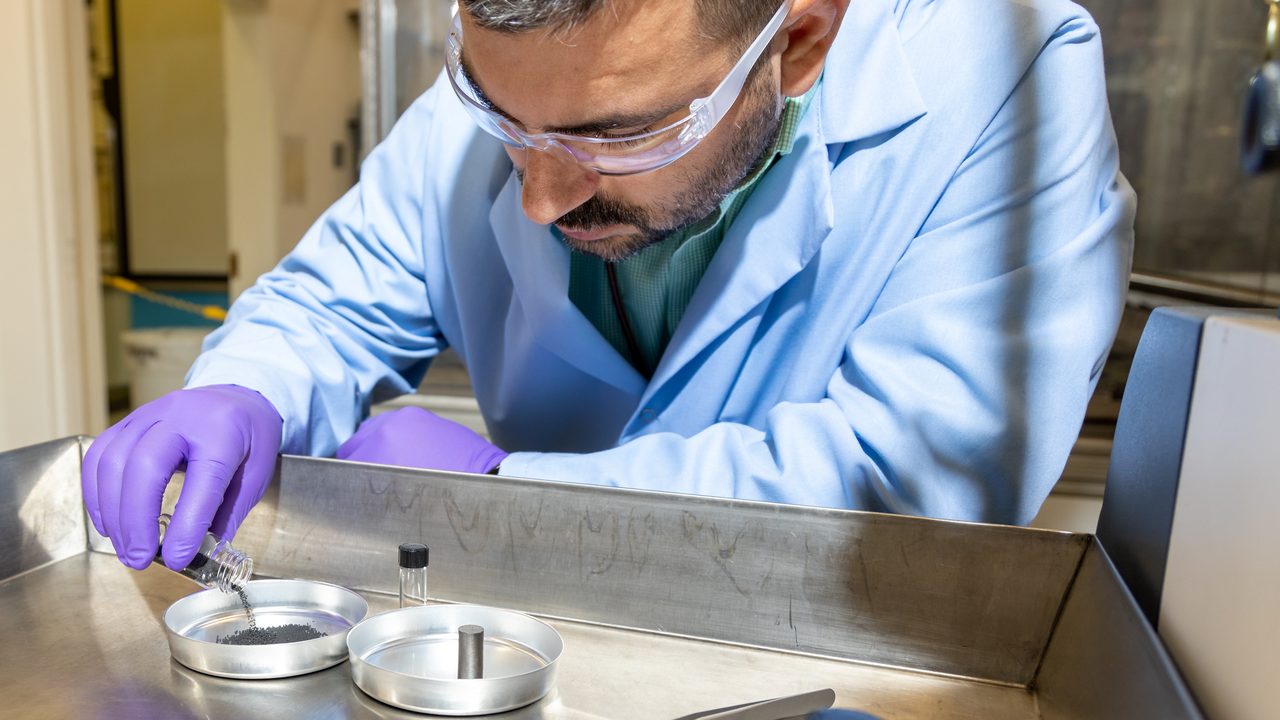 A researcher in a light blue lab coat, safety glasses and purple gloves pours dark granular particles from a small glass vial into a shallow metal dish on a steel bench.