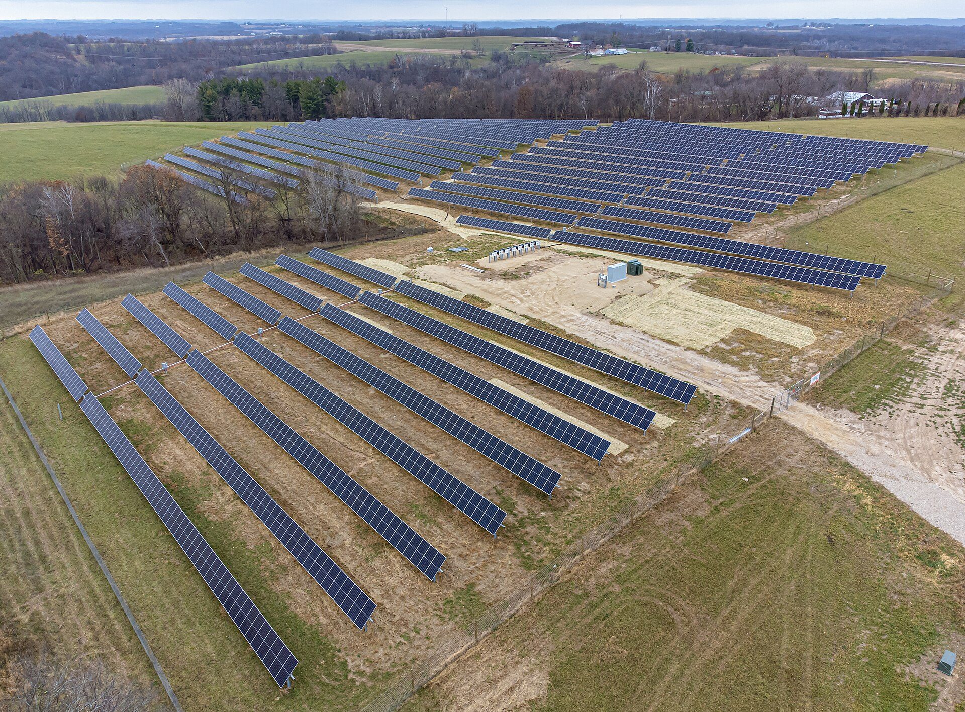 Aerial view of rows of ground-mounted solar panels on a fenced field beside bare trees and farmland