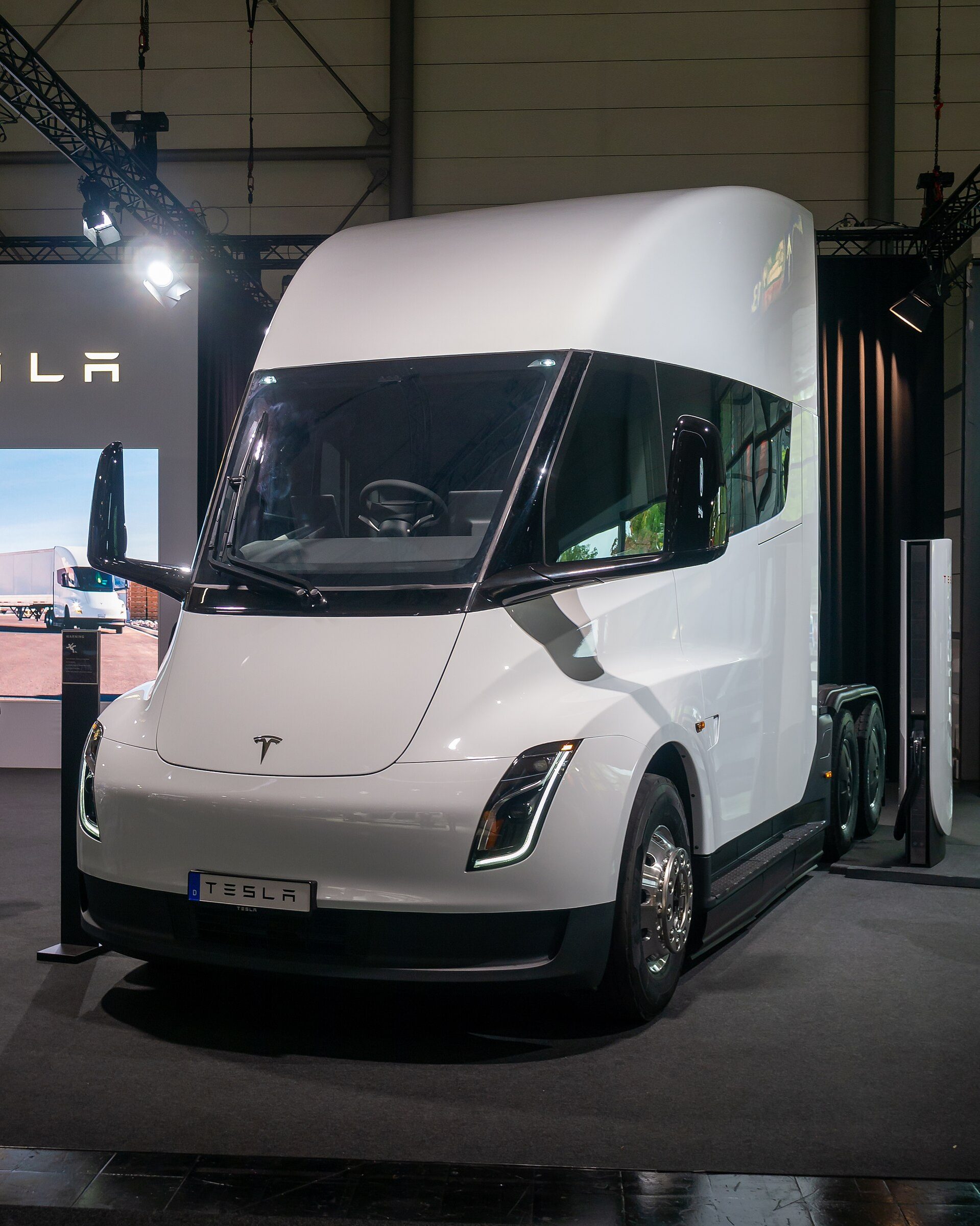 A white Tesla Semi tractor with a German show plate reading TESLA displayed indoors under stage lights at IAA Transportation 2024, with a charging post standing at its right side