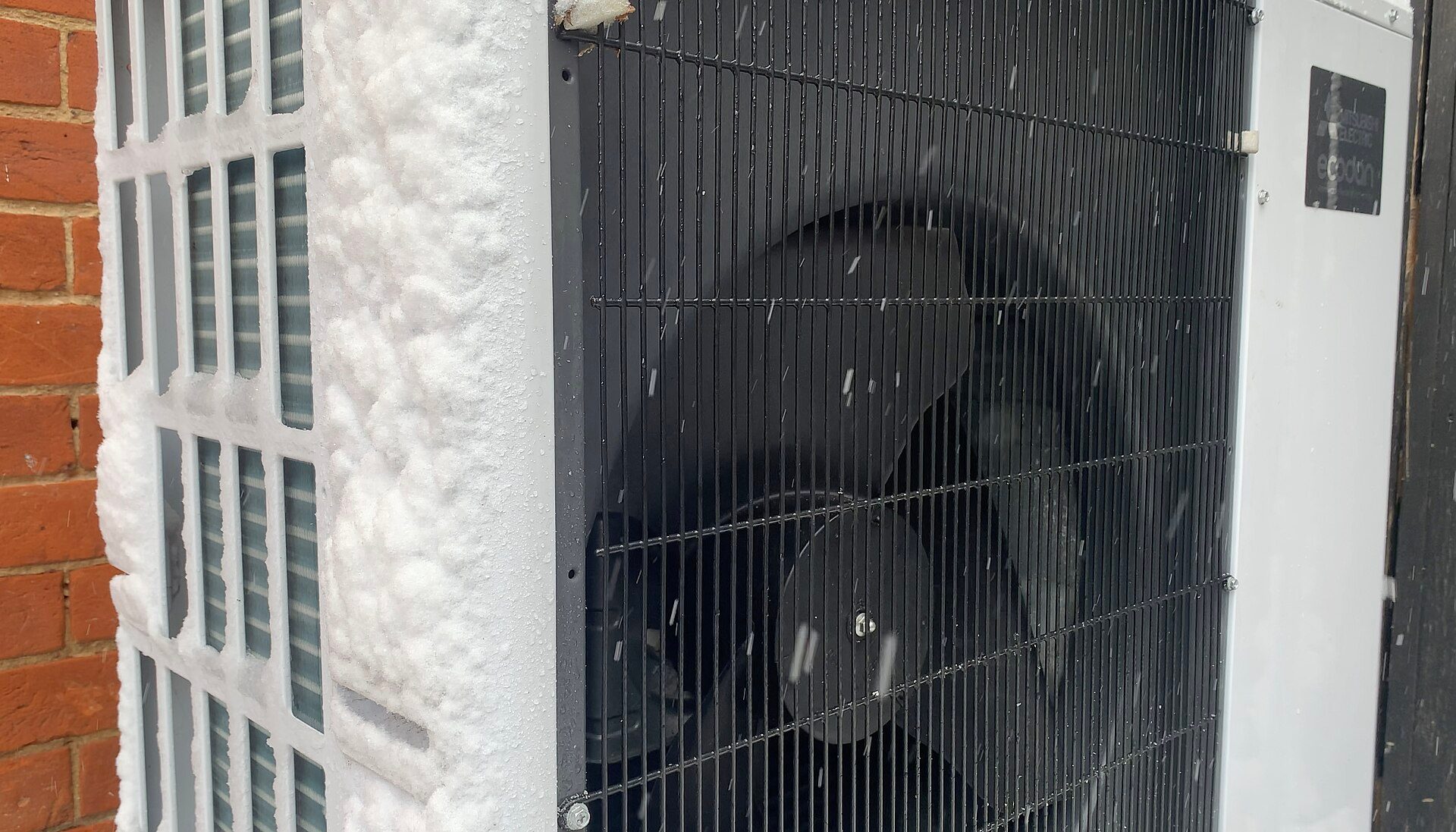 Close-up of a Mitsubishi Electric Ecodan air-source heat pump outdoor unit with snow packed against its side coil and snow falling past the black fan grille, next to a red brick wall