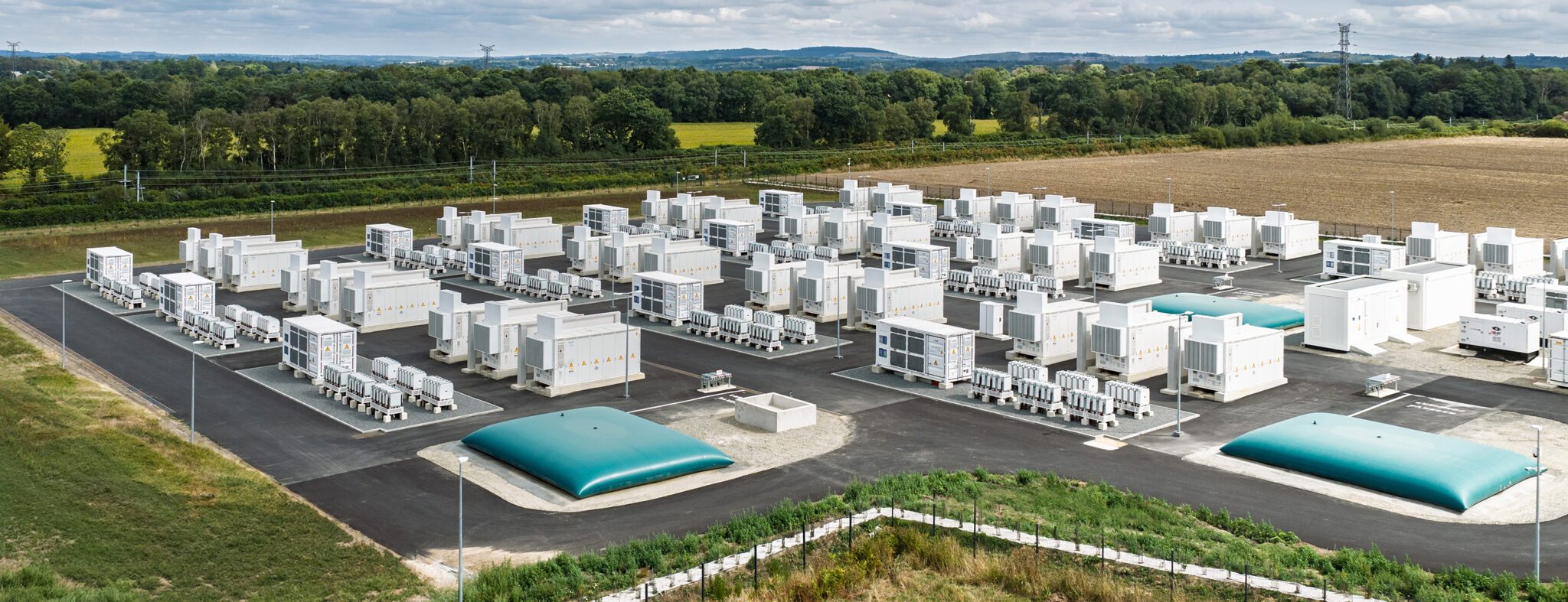 Aerial view of rows of white battery containers and smaller white equipment units on an asphalt site, with two teal domed tanks in the foreground, surrounded by fields, woodland and a power line