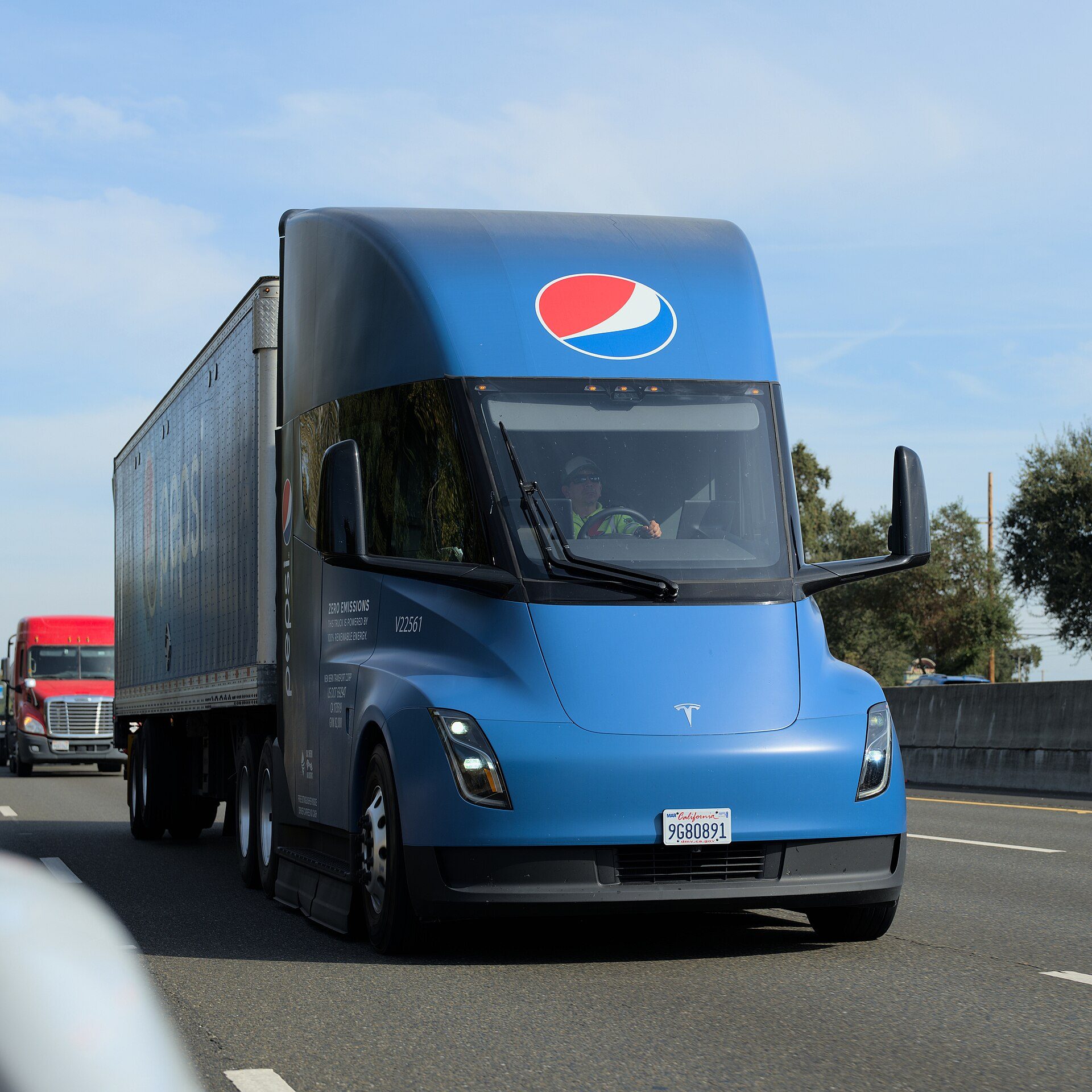 A blue Pepsi-branded Tesla Semi tractor pulling a trailer along a multi-lane highway under a clear sky, with a red conventional truck in an adjacent lane behind it