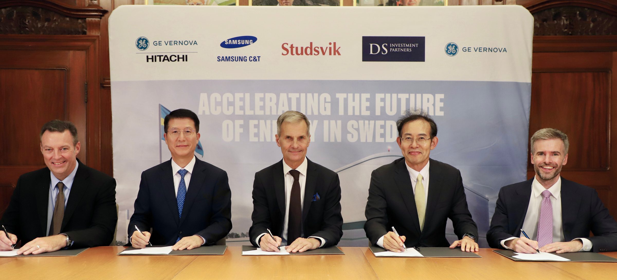 Five executives seated at a table signing documents in front of a banner carrying the GE Vernova Hitachi, Samsung C&T, Studsvik, DS Investment Partners, and GE Vernova logos
