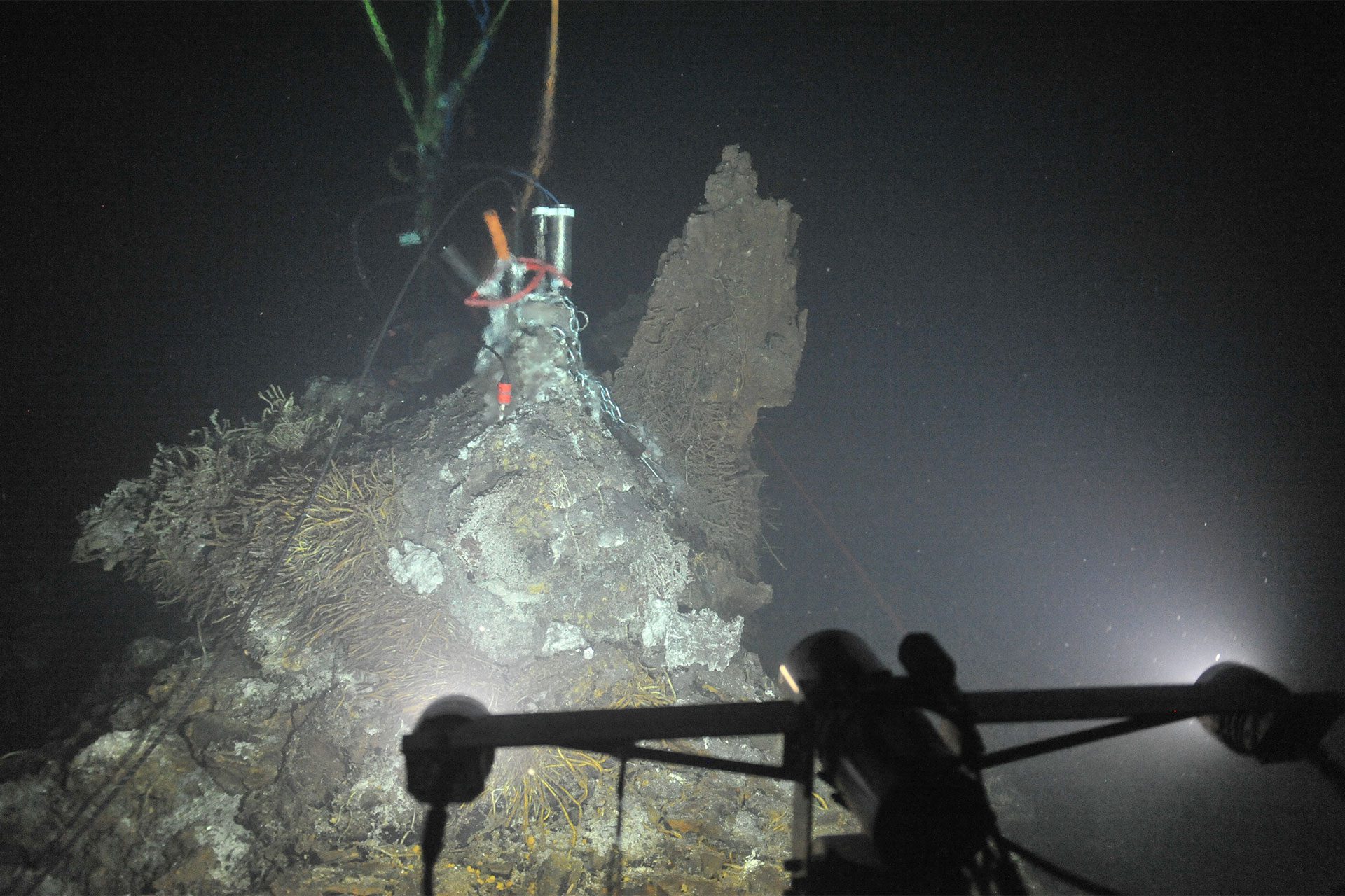 A mineral chimney at a deep-sea hydrothermal vent, crusted with pale deposits and tube-like growth, with cables and an instrument canister at its top and a lit equipment frame in the dark water in the foreground.