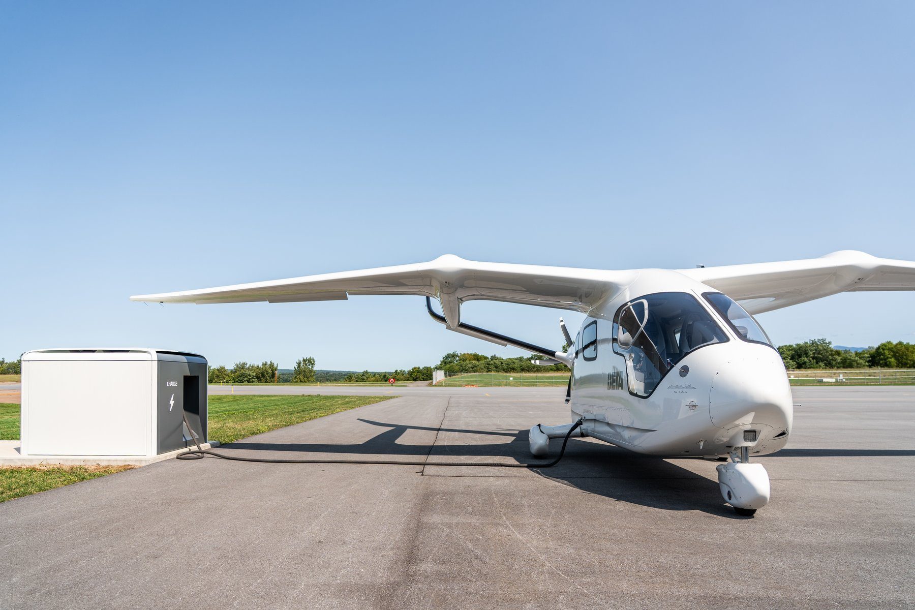 BETA's white ALIA CX300 electric aircraft parked on a paved airport apron, its nose cable plugged into a white-and-black BETA Charge Cube charger, with grass, trees and a clear blue sky behind