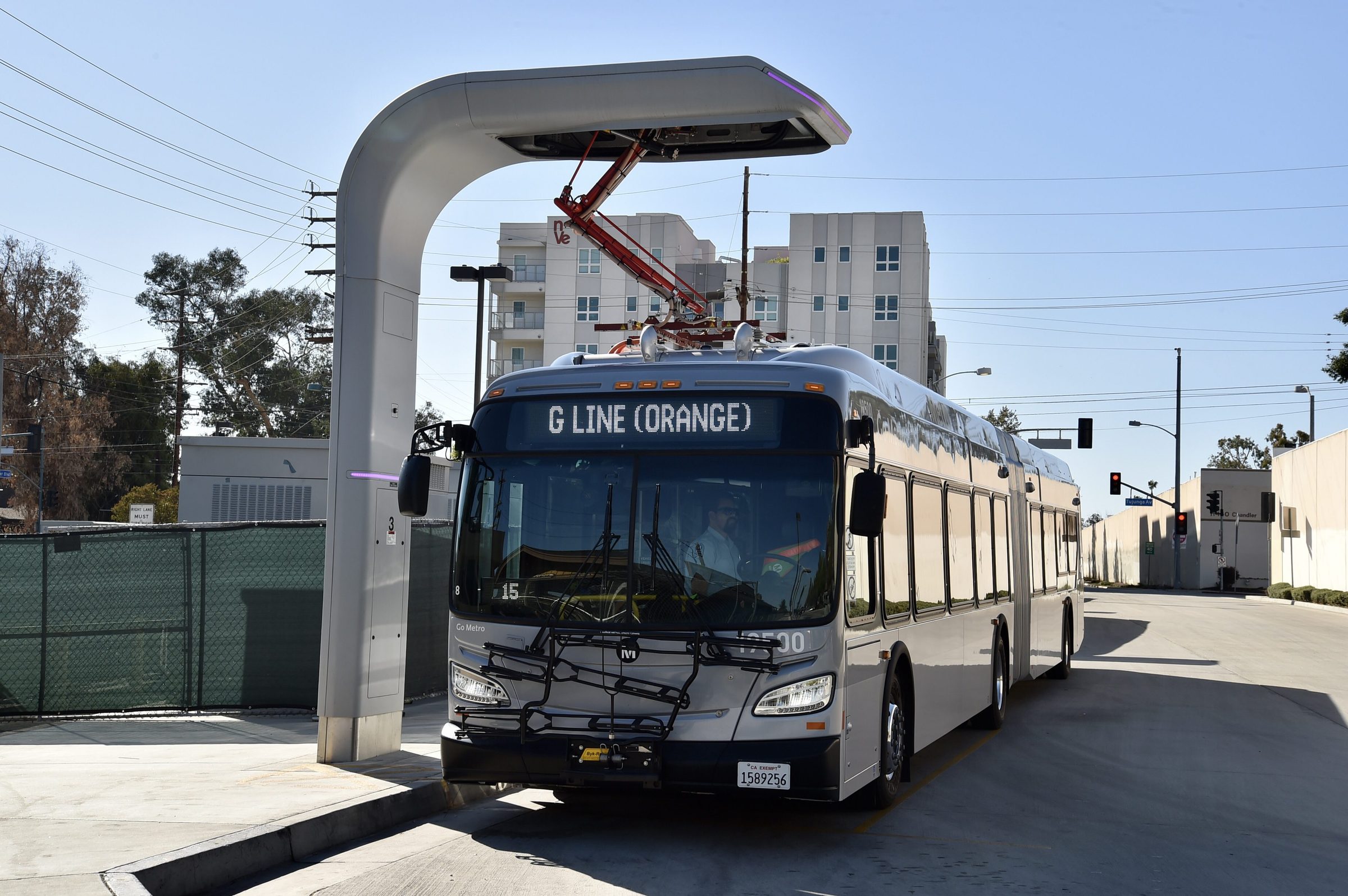 A silver Metro articulated bus with 'G LINE (ORANGE)' on its destination sign stopped at a station charger, its pantograph arm lowered onto the bus roof, a multistory building behind it