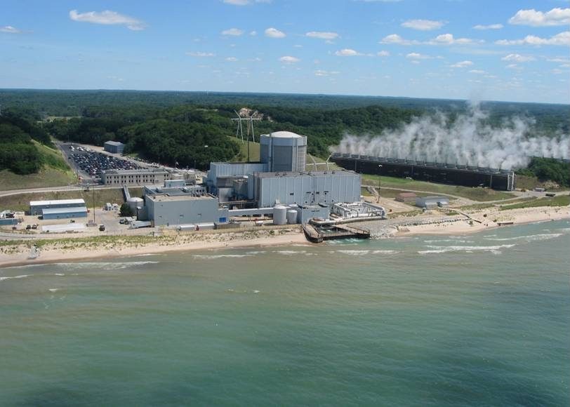 Aerial view of the Palisades nuclear plant on the Lake Michigan shore, its domed cylindrical containment building near the beach and a long bank of cooling towers venting steam behind it
