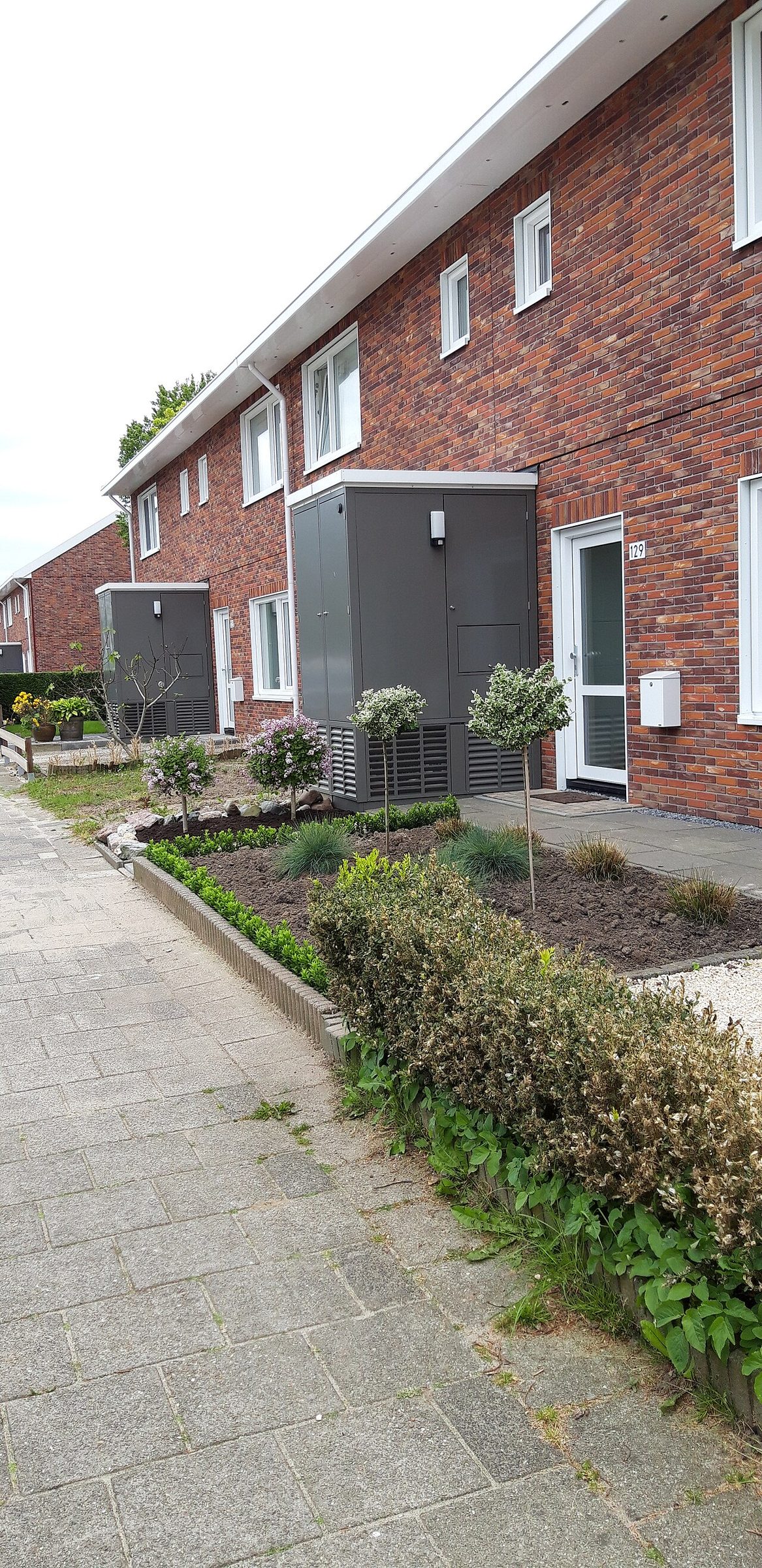 Grey heat pump cabinets standing in front of red-brick terraced houses, beside small planted front gardens and a paved sidewalk