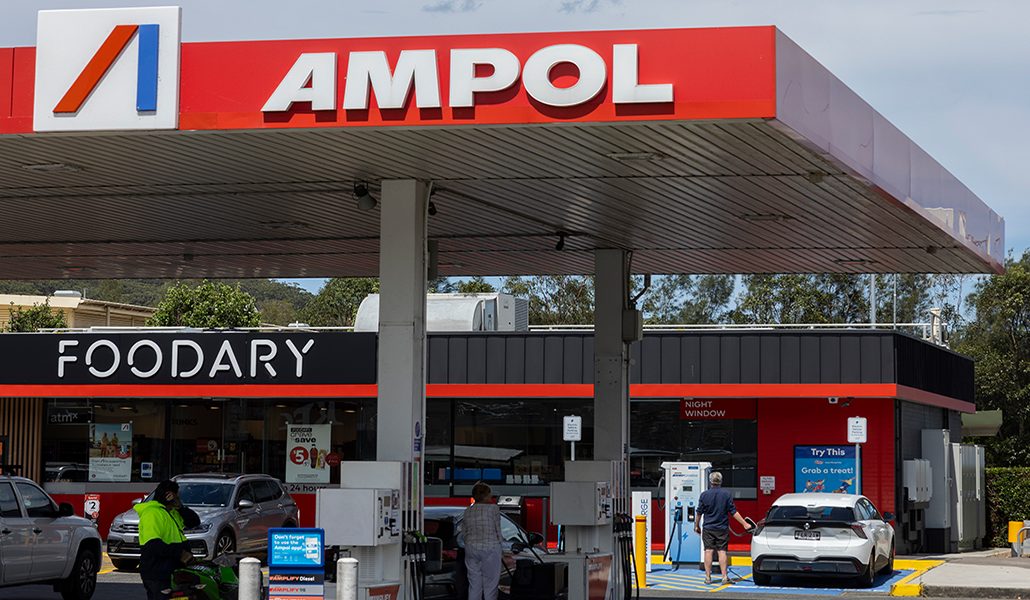 An Ampol service station with a Foodary store under a red Ampol canopy, fuel pumps in the foreground and a man standing at an electric vehicle charger beside a white hatchback on the right