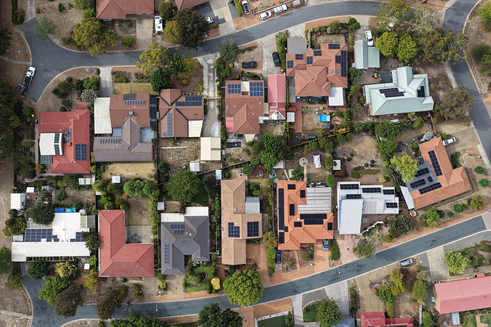Aerial view straight down over a Canberra suburb: houses with terracotta, grey and pale metal roofs, many carrying rectangular solar panel arrays, along curving streets with parked cars, trees and dry lawns.