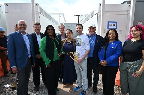 Nine people pose between two rows of white battery enclosures at NineDot's Hunts Point site, two of them holding a large pair of gold ceremonial scissors, with a worker in an orange vest partly visible at the left edge and a sign reading LOT 641 on one enclosure