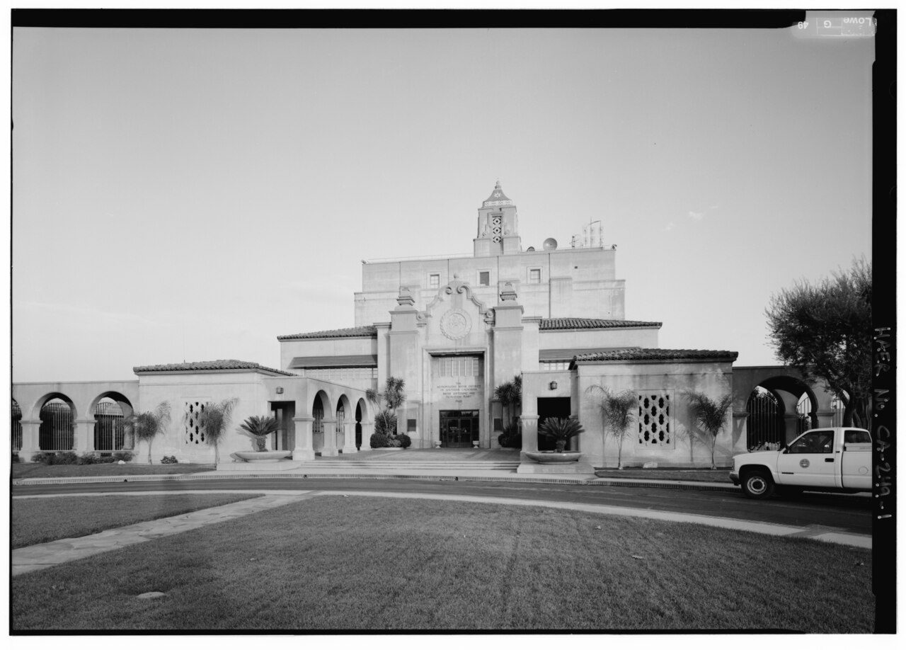 Black-and-white 1998 Historic American Engineering Record photograph of the west face of the F.E. Weymouth plant's main building in La Verne: a Spanish-style facade with a central tower, arched colonnades on each side, palm trees, a lawn in the foreground and a white pickup truck parked at right
