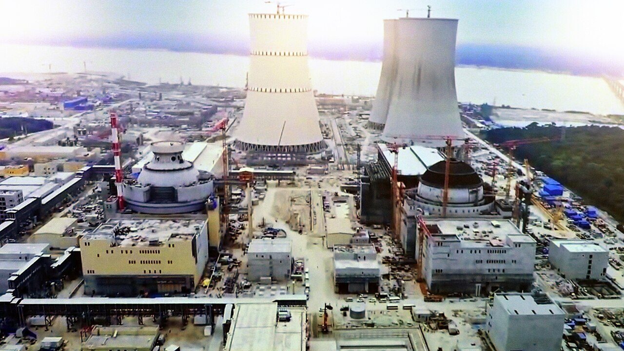 Aerial view of the Rooppur nuclear plant under construction: two domed reactor buildings ringed by cranes, three cooling towers, and a wide river behind.