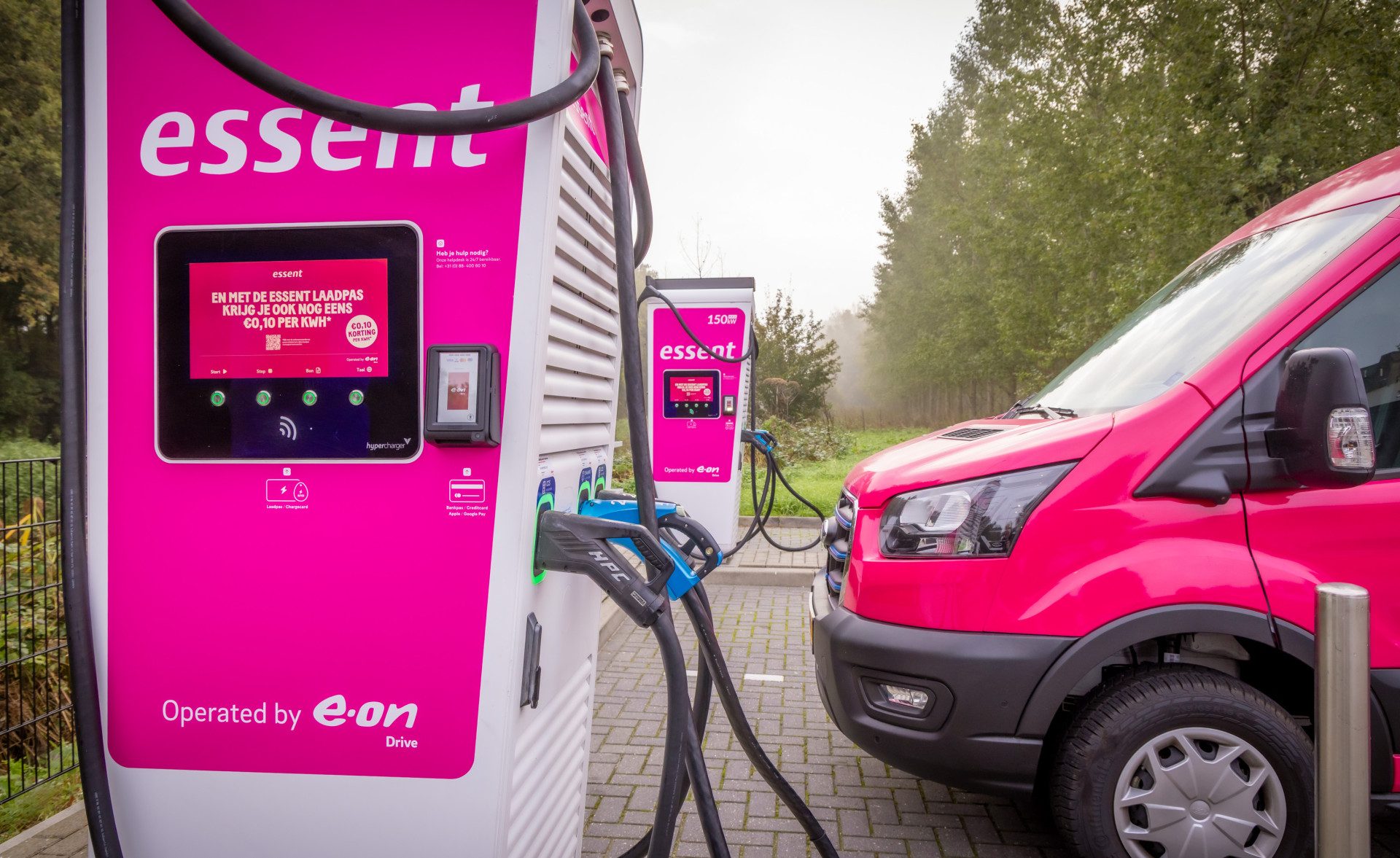 Pink Essent-branded fast charger marked Operated by E.ON Drive with its two connectors holstered at the side, a second Essent unit labelled 150 kW behind it and a pink van parked alongside, not plugged in