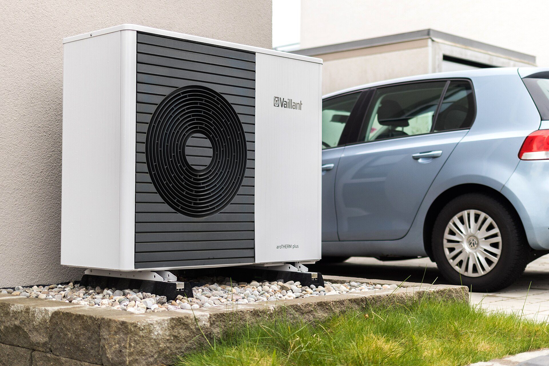 A white Vaillant air-to-water heat pump outdoor unit standing on rubber feet over a gravel bed beside a rendered house wall, with a light blue hatchback parked behind it