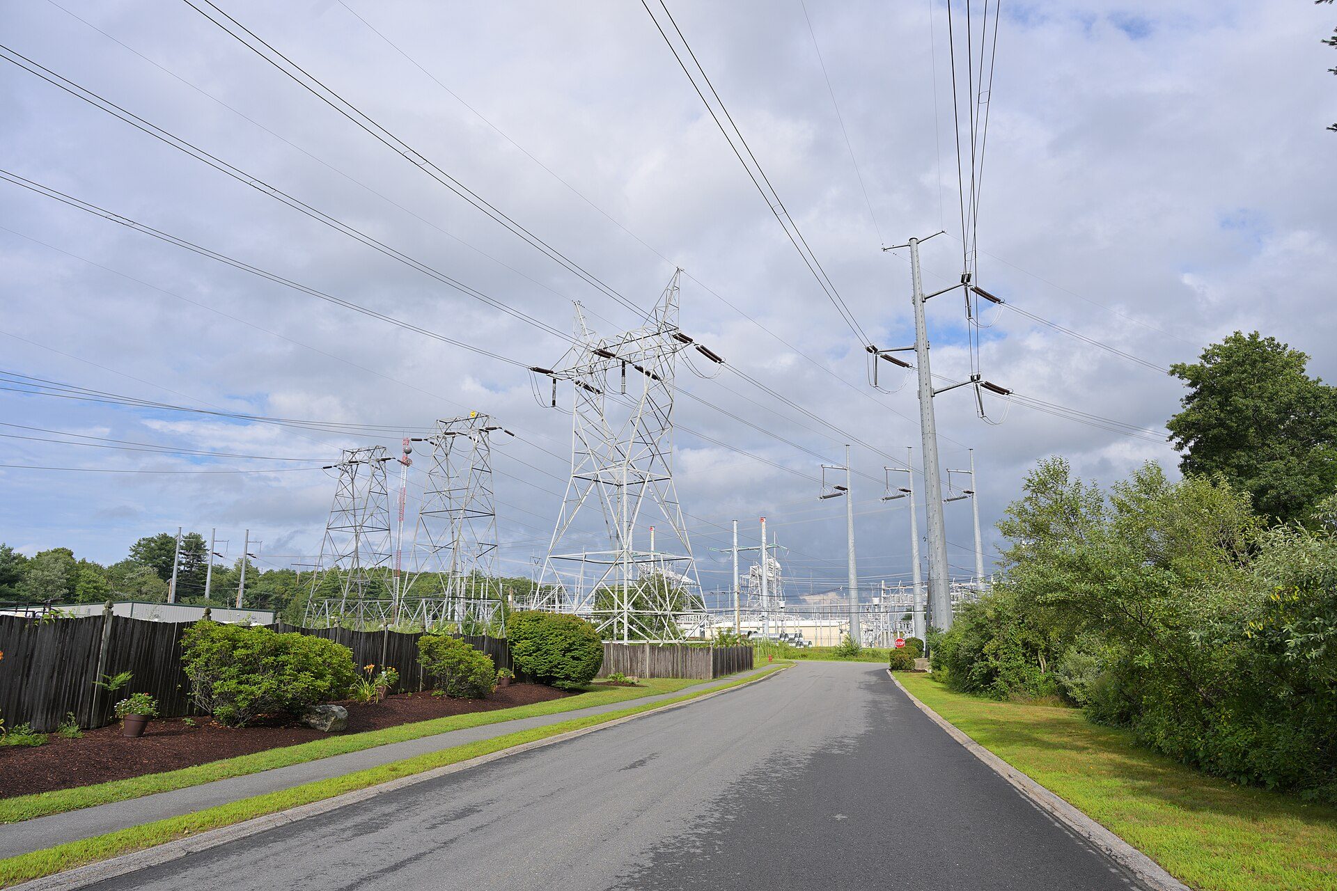 Steel lattice towers and tall steel poles carry high-voltage lines over a paved road toward substation equipment at Sandy Pond in Ayer, Massachusetts