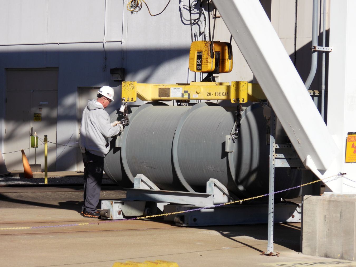 A worker in a white hard hat and grey hooded sweatshirt attaches a chain hook to a horizontal grey steel cylinder resting on a cradle beneath a yellow 20-ton spreader bar, outside a light-coloured industrial building in sunlight