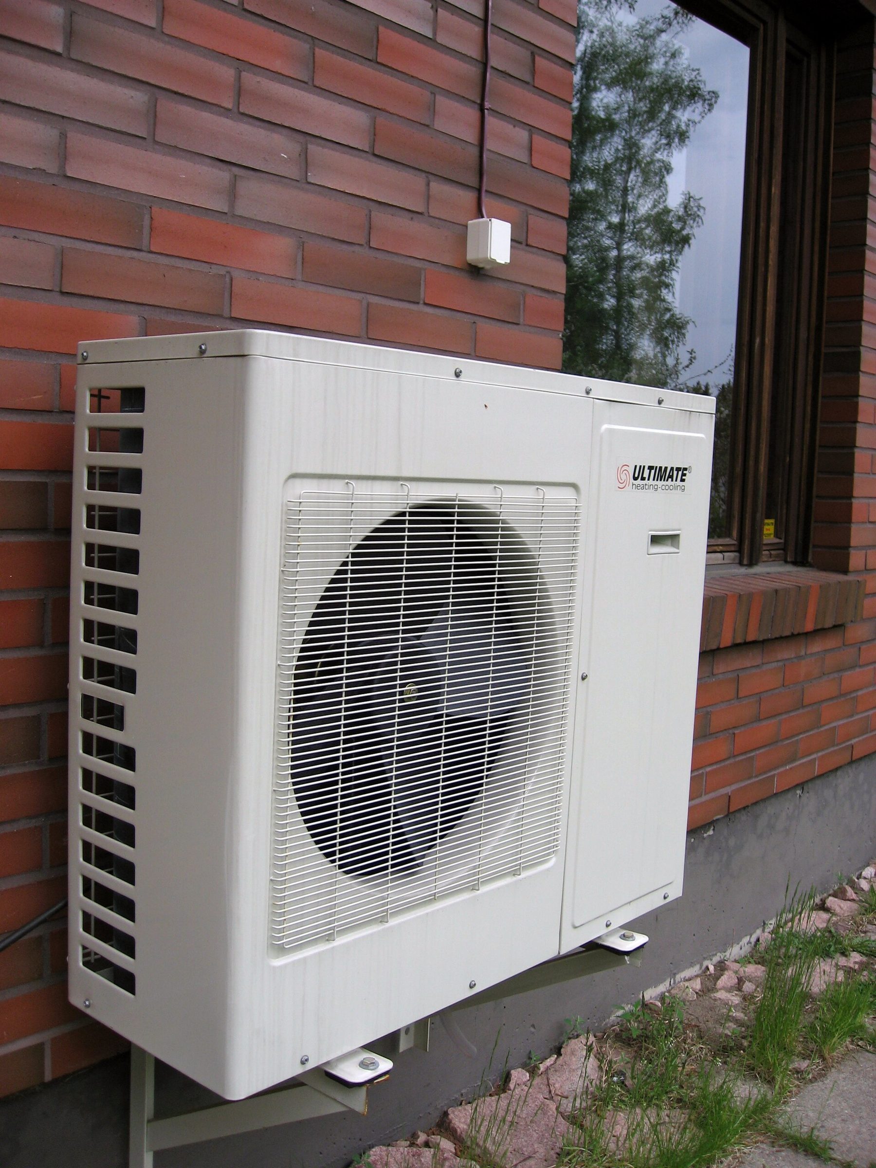 White outdoor unit of an air-source heat pump mounted on brackets against a red brick wall beside a window, with grass and stones below.