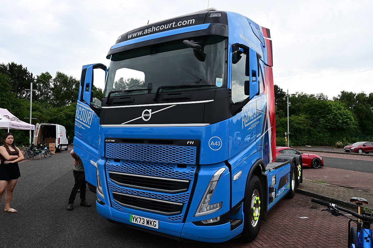 Blue Volvo FH Electric tractor unit in Ashcourt Group livery parked on a car park with one cab door open, a white van and a gazebo behind it