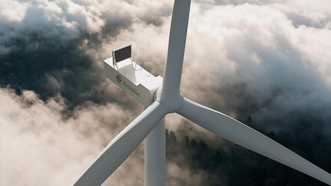 An Envision wind turbine nacelle and three-blade rotor seen from above, with cloud banks and forested hills below
